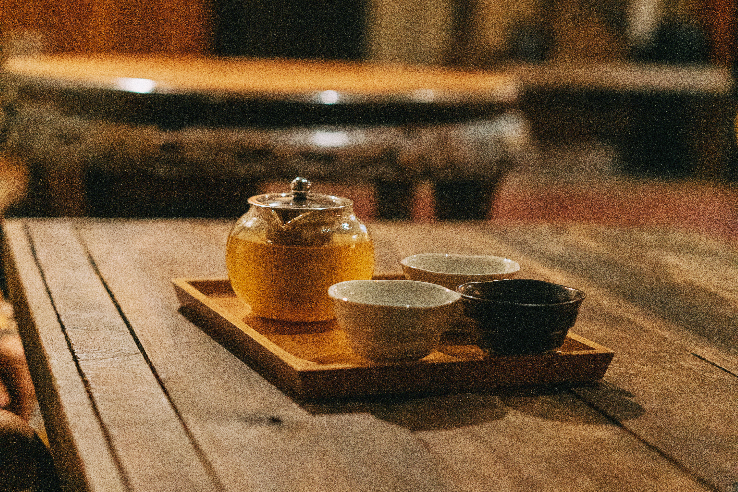 The image shows a glass teapot with a metal lid, filled with yellow tea, and three cups on a wooden tray placed on a rustic wooden table in a cozy, dimly lit setting.