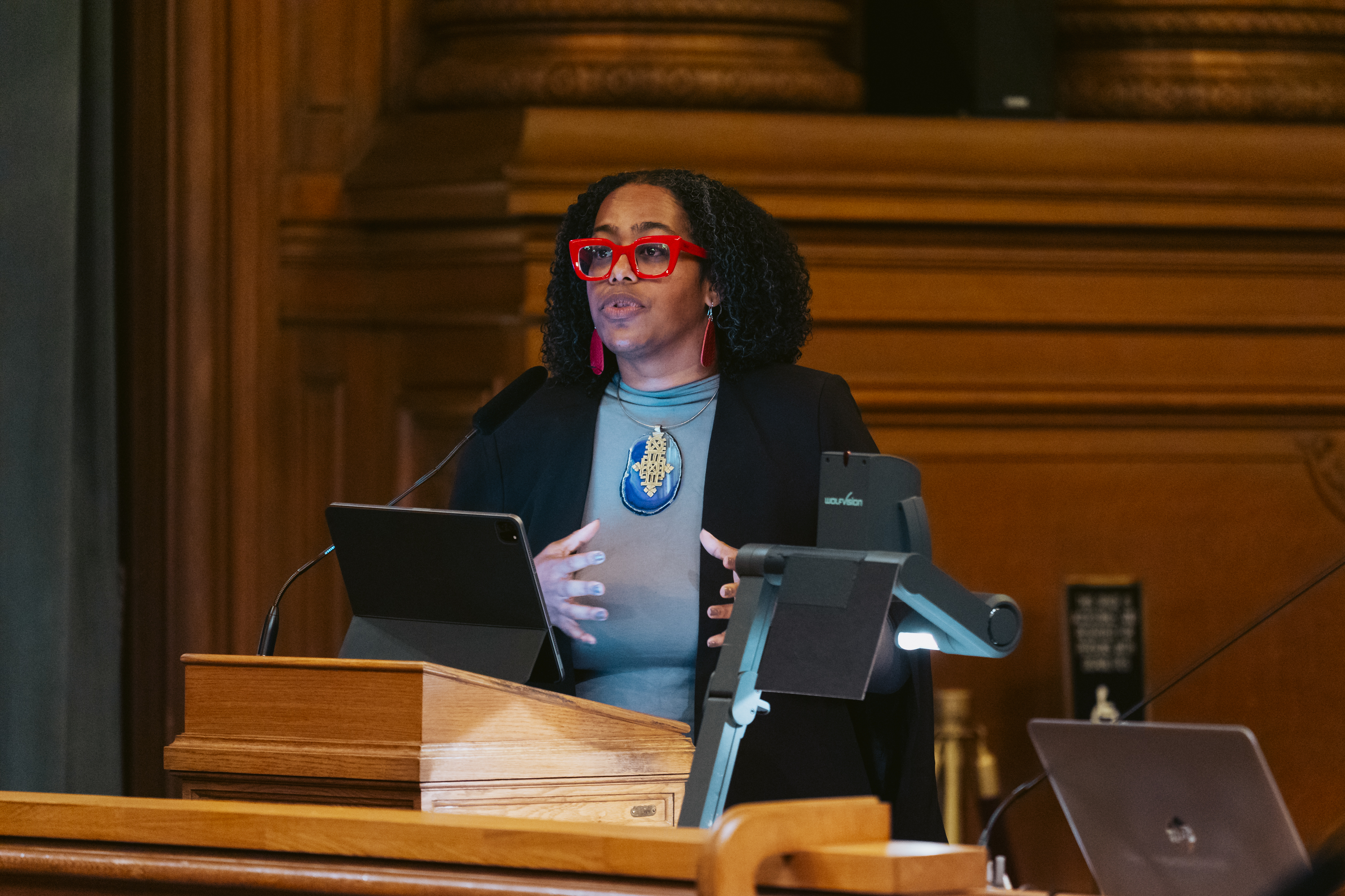 A person with curly hair and red glasses speaks at a podium, wearing a blazer and a large pendant, surrounded by wooden décor and some electronic devices.