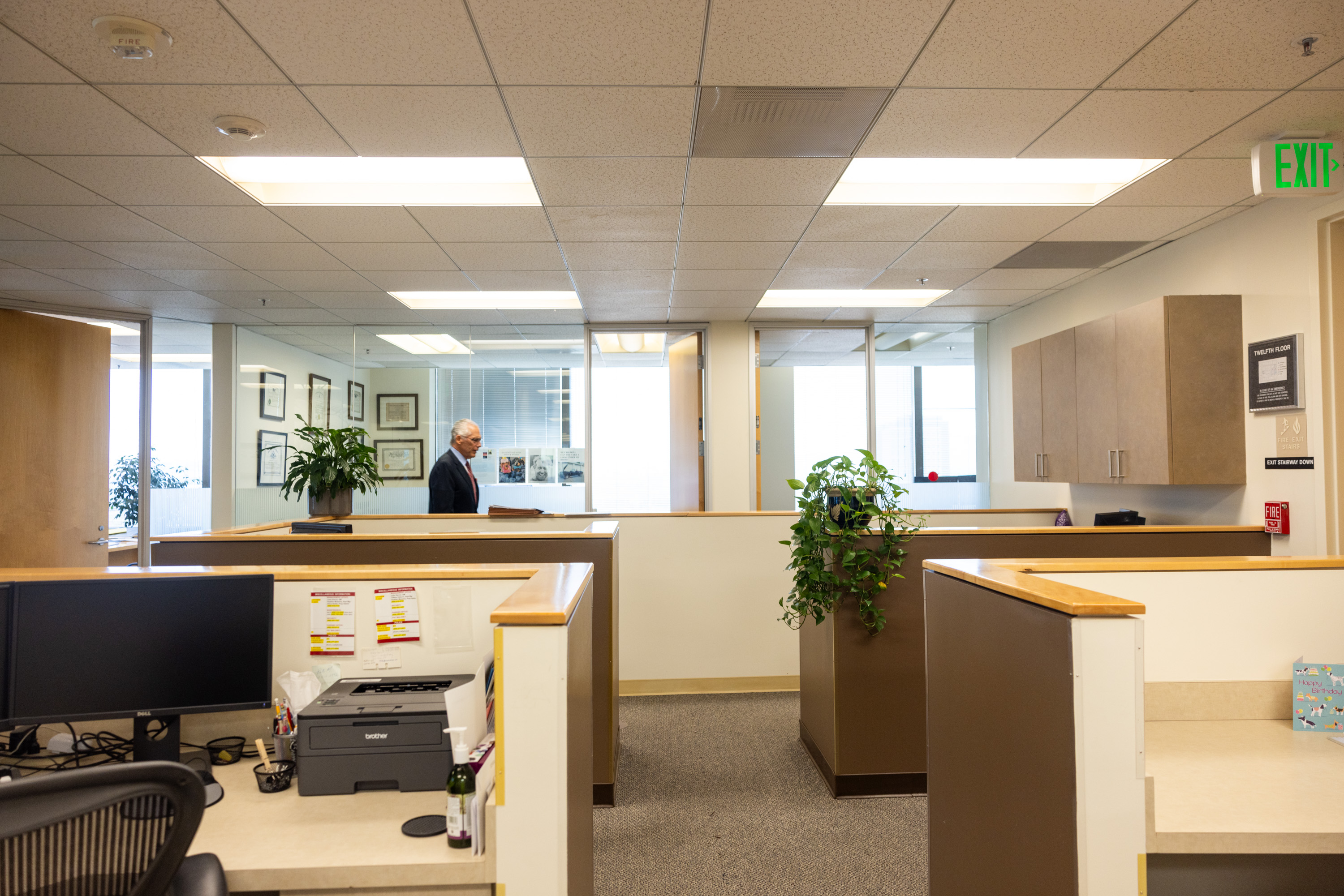 The image shows a tidy office with cubicles, plants, framed pictures, and a man walking. There's an EXIT sign above a door, and office equipment/desk supplies are visible.