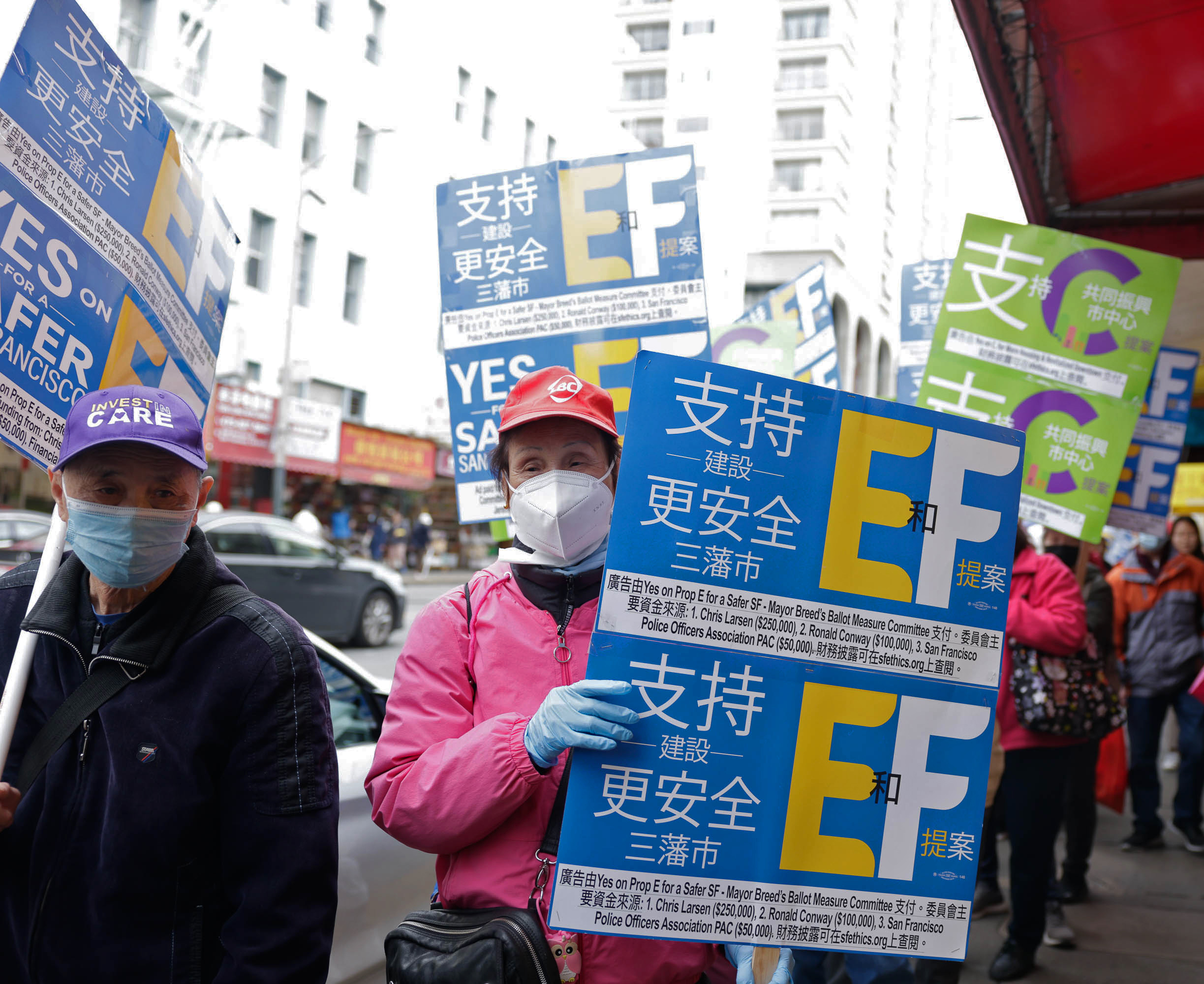 People are holding blue and green signs with &quot;EF&quot; in a city street, advocating for a proposition, wearing masks and gloves.