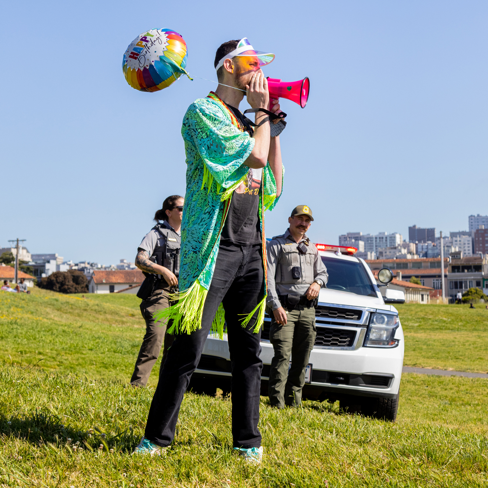 A man in a party hat and colorful shawl speaks into a megaphone at a sunny park, flanked by uniformed officers near a patrol car.