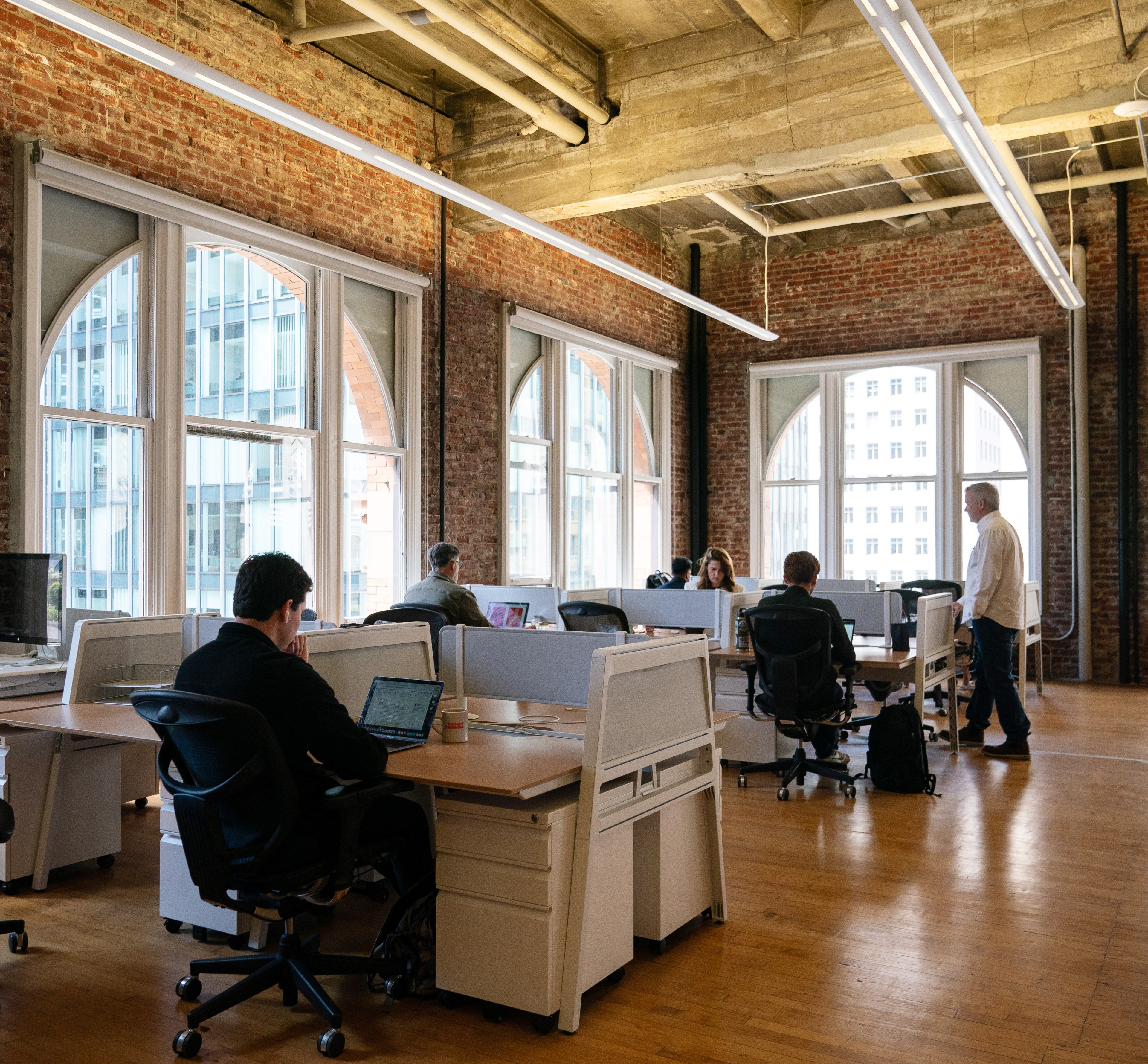 An open-plan office with exposed brick walls and large arched windows; several people are working at desks with computers, and one person is standing and talking.