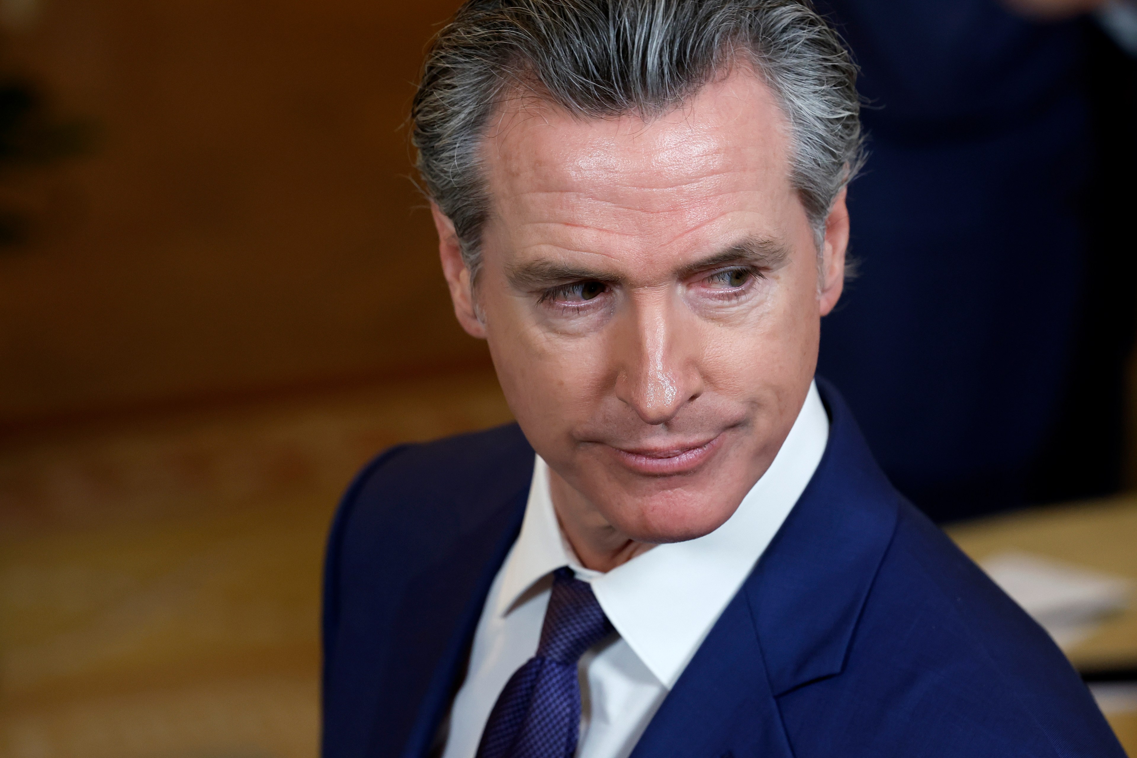 A man in a blue suit and tie looks to his left with a serious expression, set against a blurred, warm-toned background.