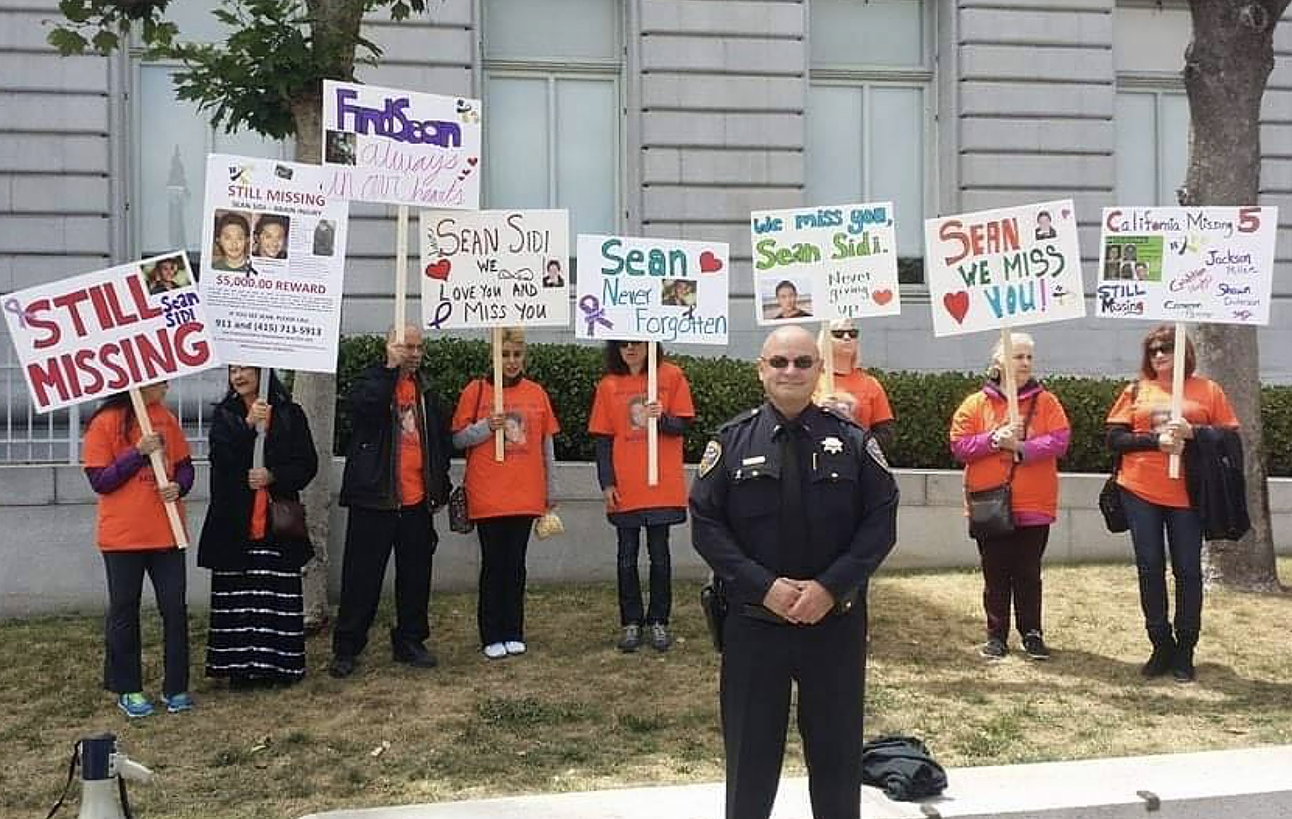 People hold signs seeking information about a missing person called Sean, as a uniformed officer stands in the foreground.
