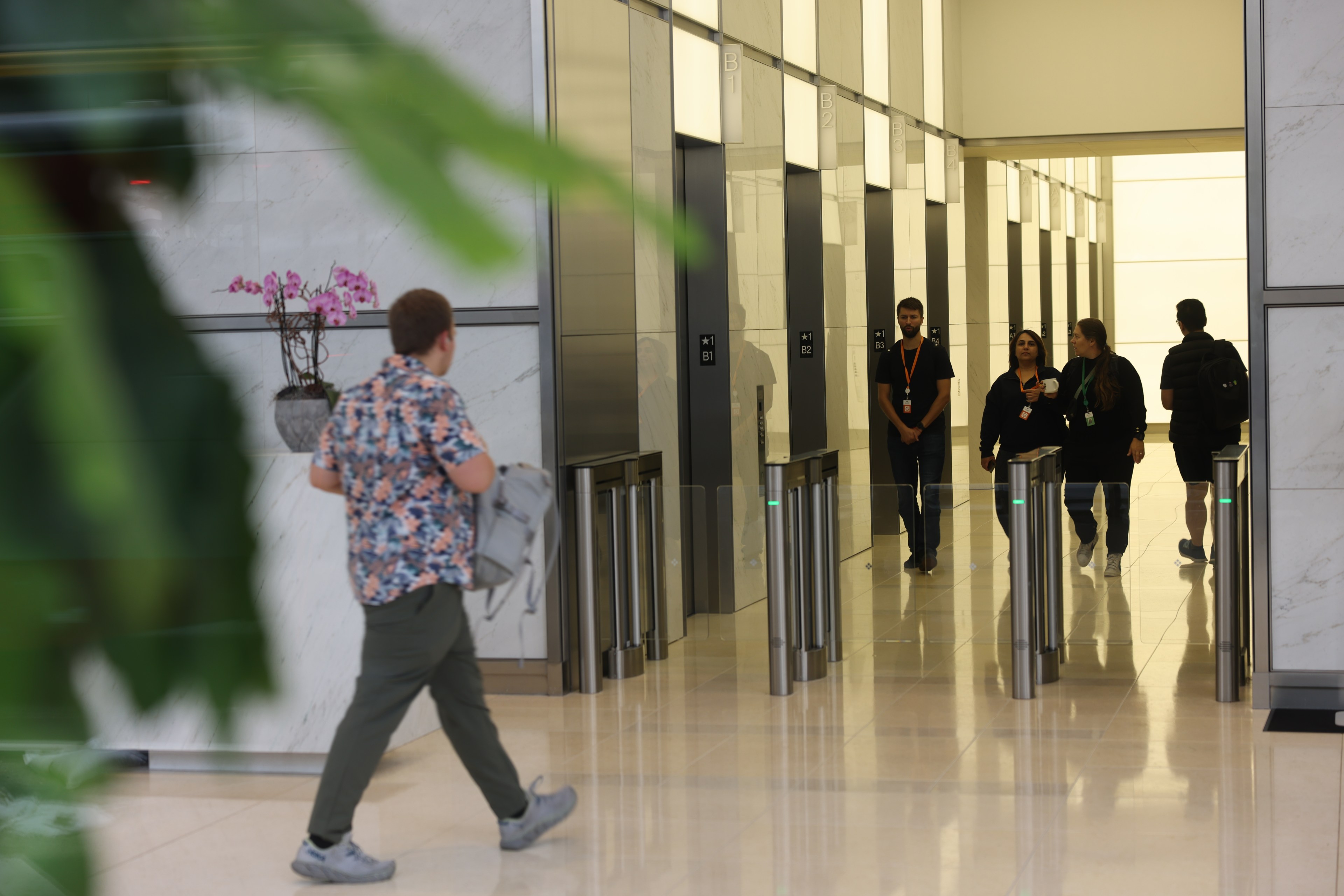 A man in a floral shirt walks towards a bank of elevators, while four other people stand or walk near the elevators in a modern, well-lit lobby with a large plant in the foreground.