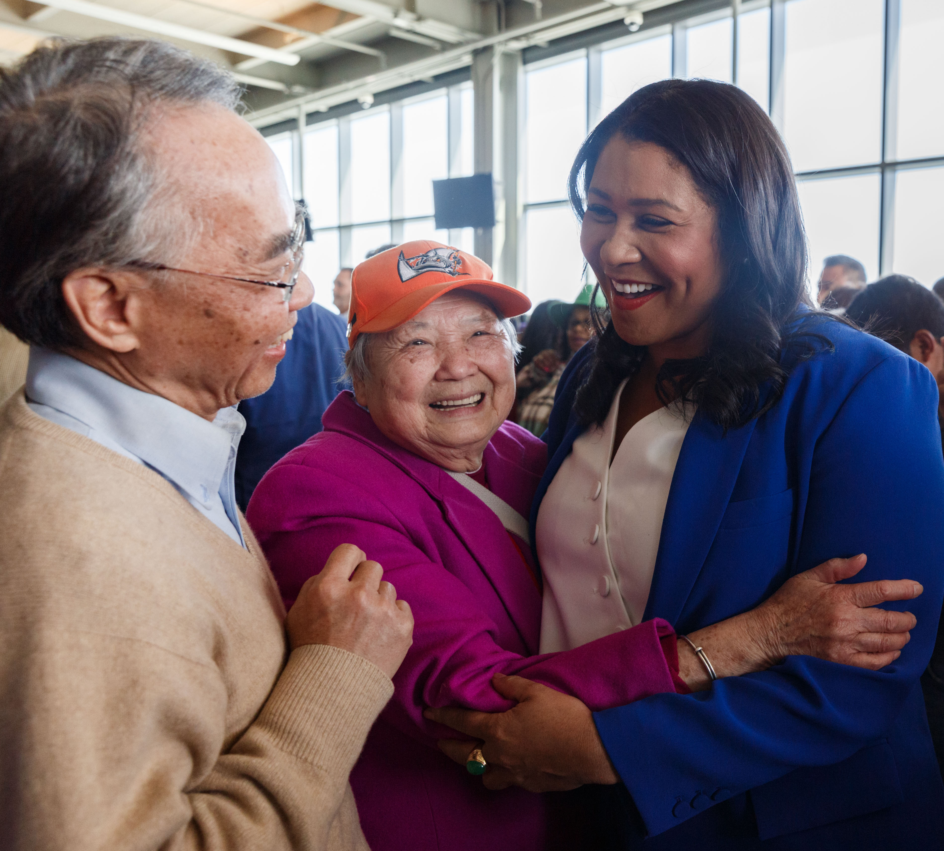 An elderly Asian couple shares a joyful moment with a smiling woman in a blue blazer, inside a bright room with large windows.