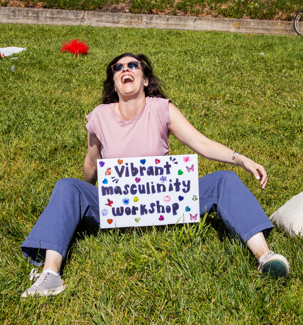 A joyful woman sits on grass behind a sign that reads &quot;Vibrant masculinity workshop&quot; that's adorned with colorful stickers.