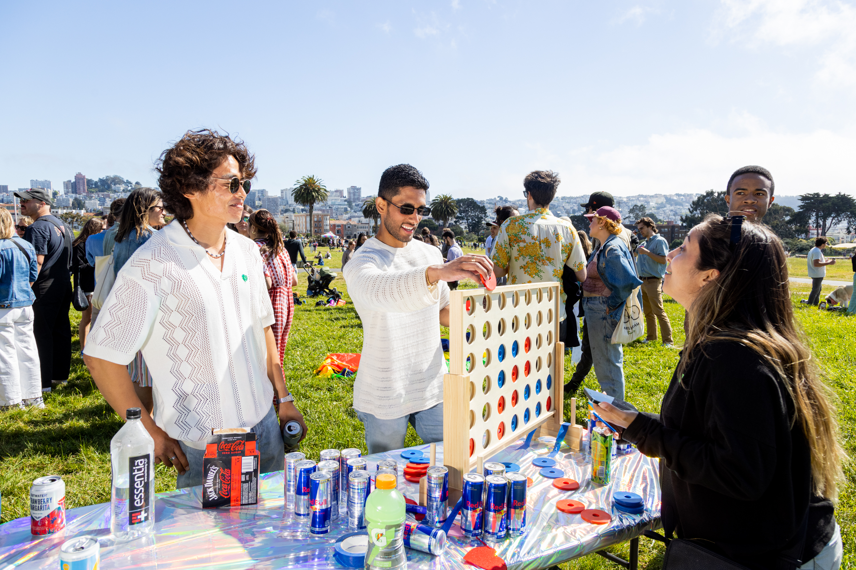 People play a Connect Four game at a sunny park gathering, with a city skyline in the background. Drinks and snacks are on the table.