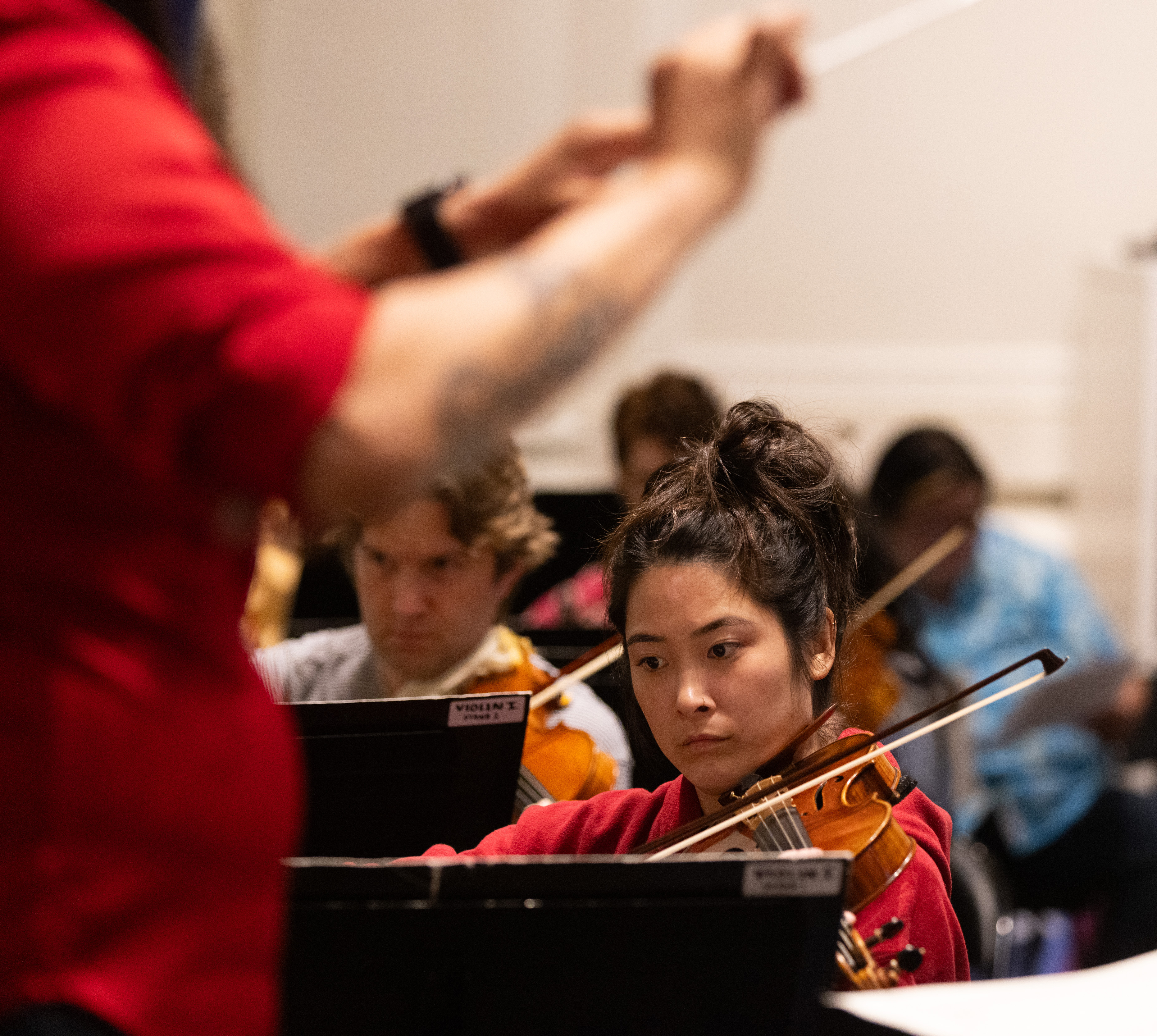 A focused woman plays the violin in an orchestra rehearsal, with a blurred conductor's arm in the foreground and other musicians in the background.