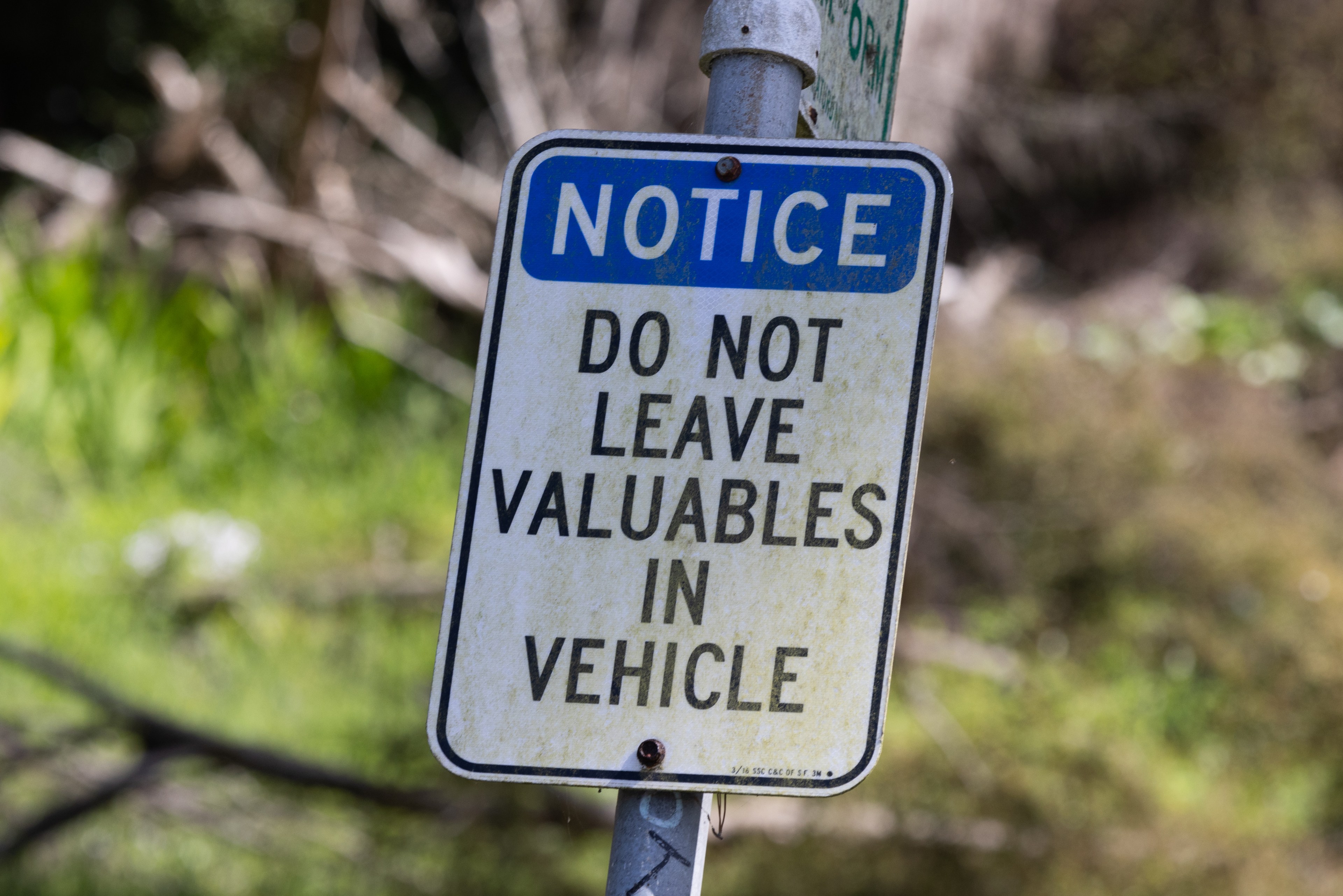 A weathered warning sign attached to a pole reads, &quot;NOTICE DO NOT LEAVE VALUABLES IN VEHICLE,&quot; set against a blurred natural background.