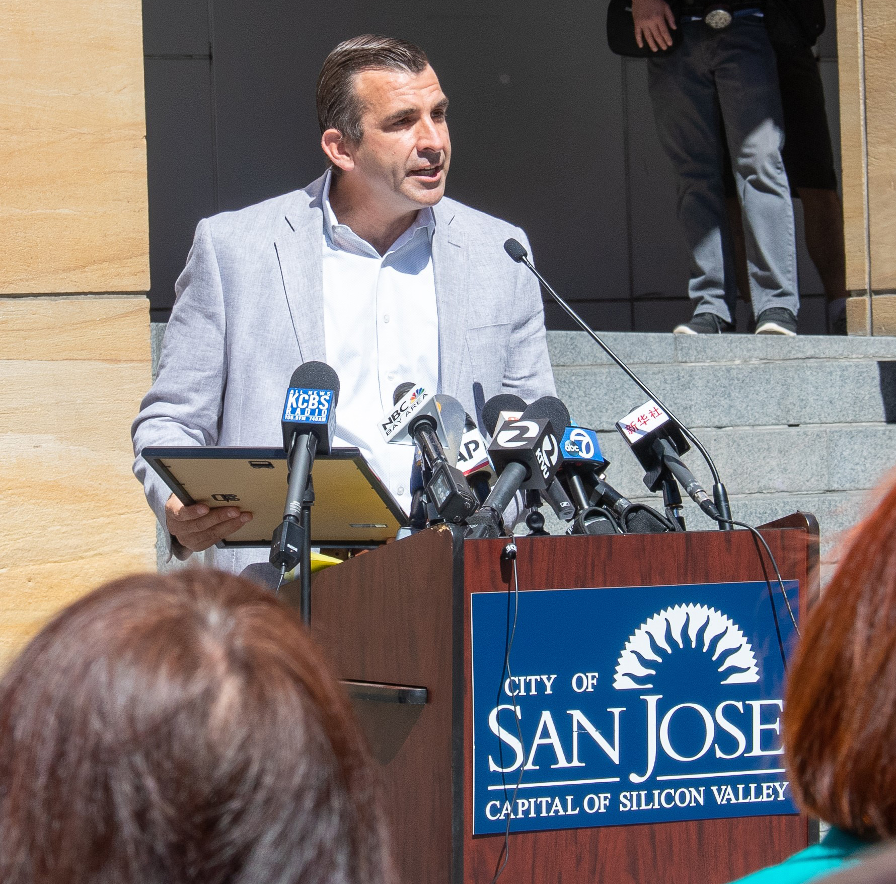 A man in a light gray jacket speaks at a podium surrounded by microphones. The podium has a sign that reads &quot;City of San Jose, Capital of Silicon Valley.&quot;