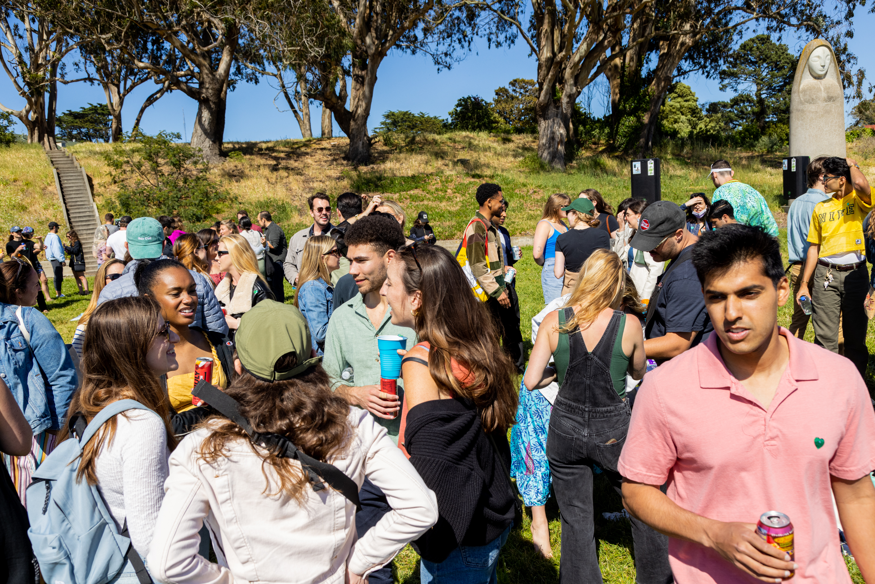 A diverse crowd of people gathered in a grassy area socialize and enjoy a sunny day outdoors. Some hold drinks.