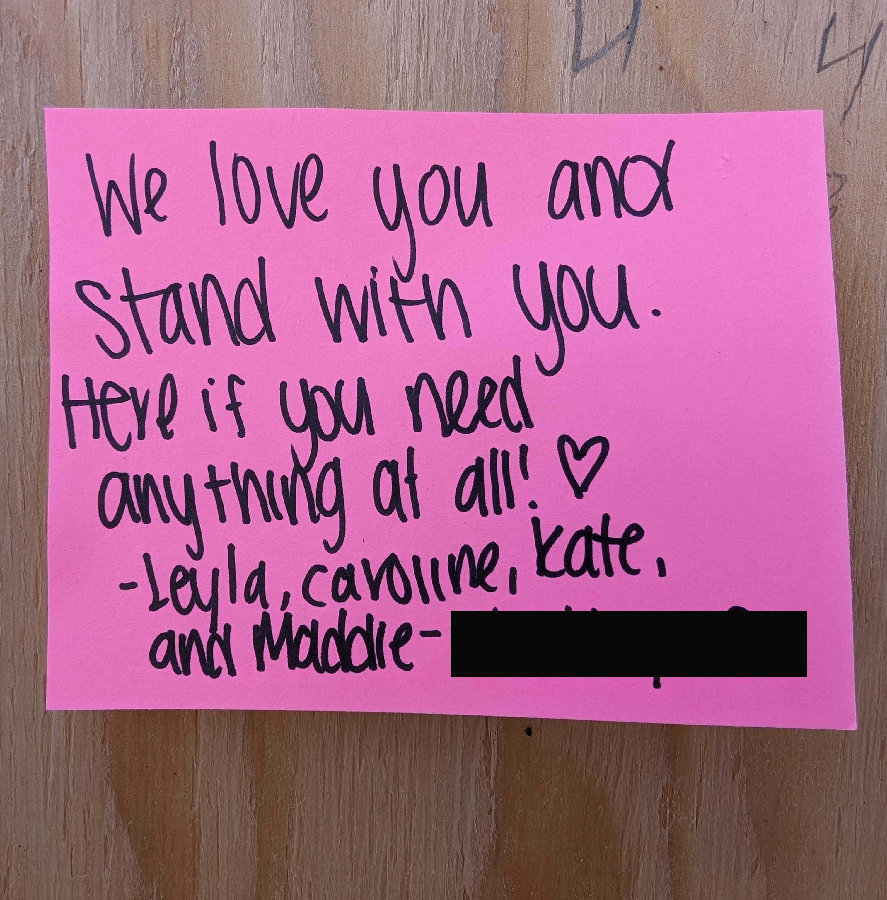 The image shows a pink note with black handwritten text that reads: &quot;We love you and stand with you. Here if you need anything at all! ♥ -Leyla, Caroline, Kate and Maddie&quot;.
