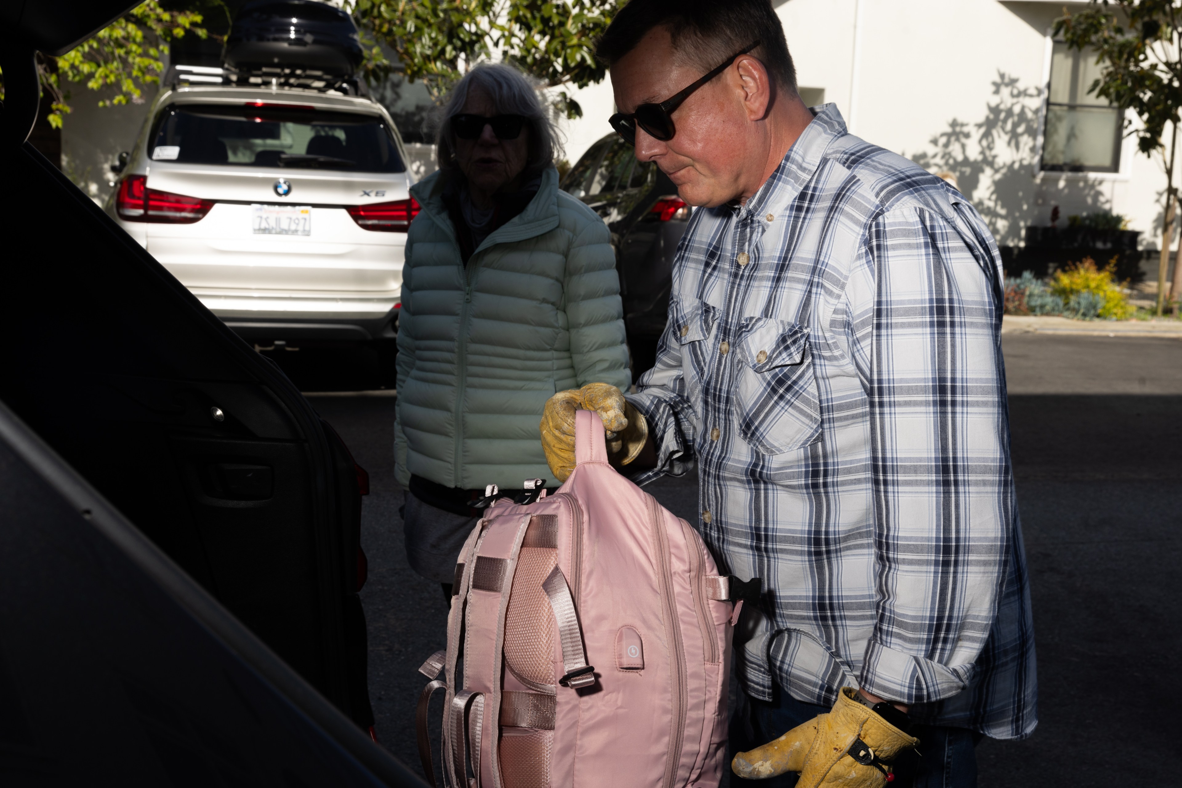 A man in a checked shirt loads a pink backpack into a car trunk; an older woman in a light green jacket observes.