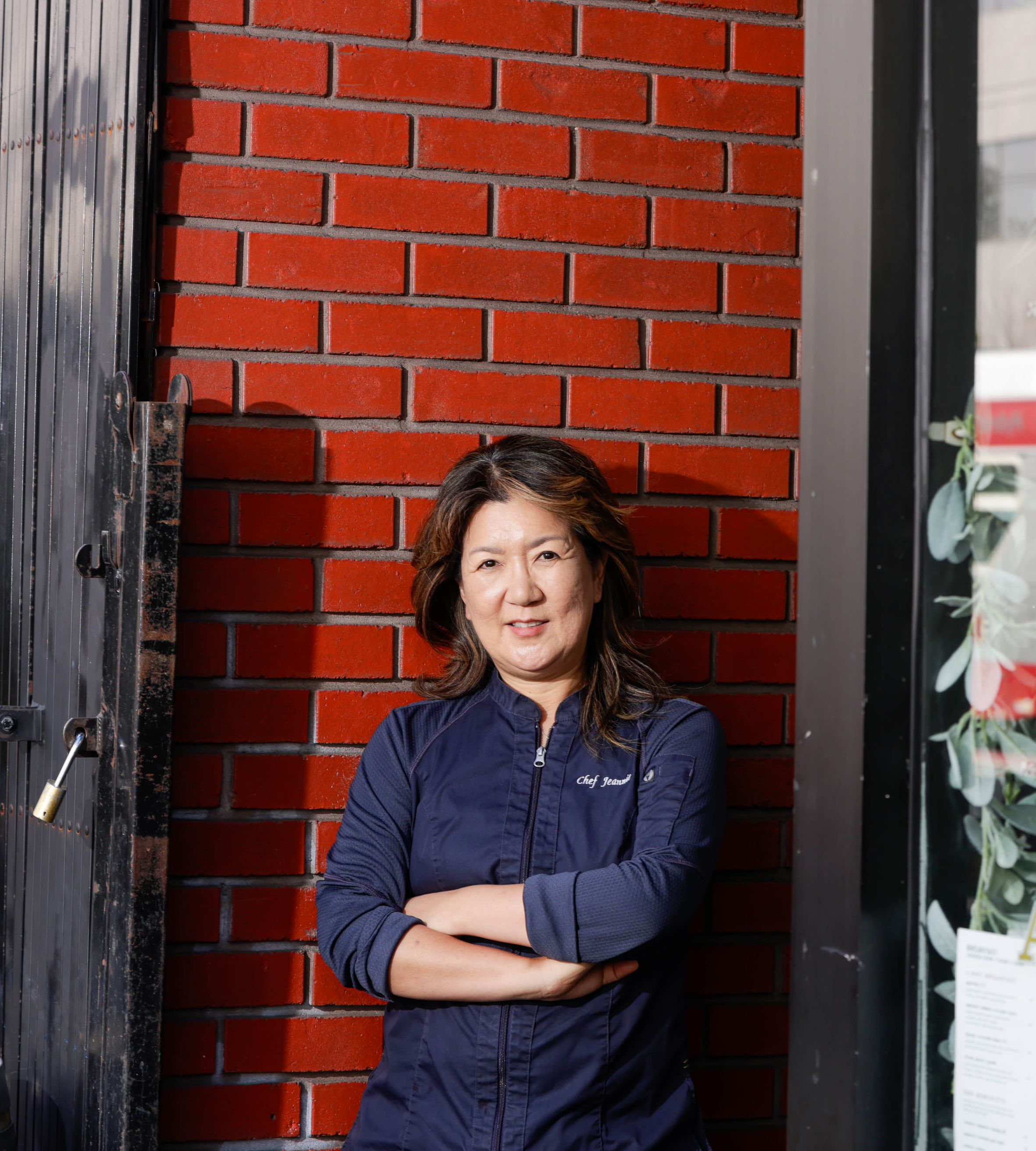 A woman in a chef's jacket stands with arms crossed by a brick wall, near a door.