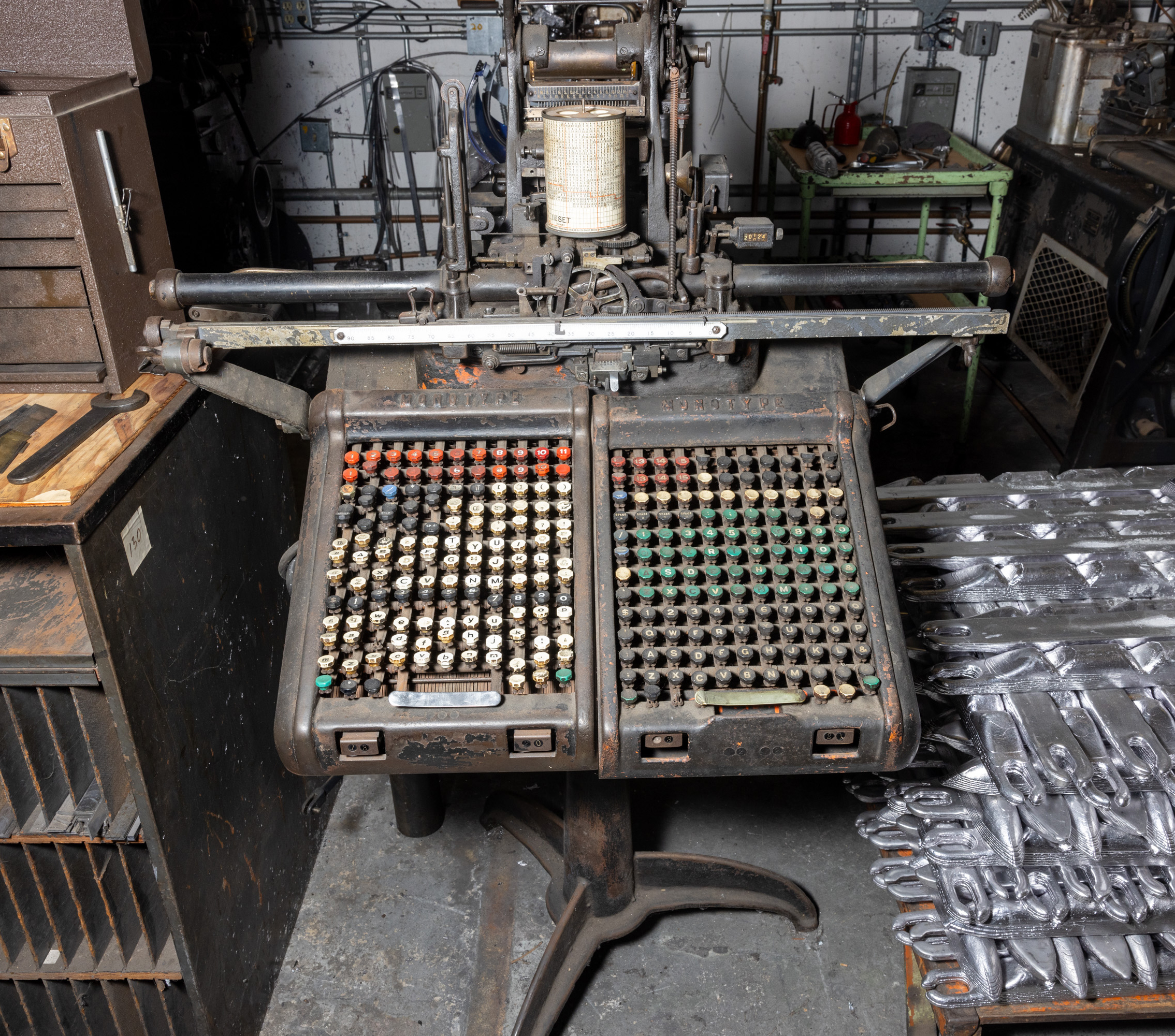 A vintage printing press in a cluttered workshop, with an old, worn-out typesetting machine featuring multiple colorful keys in the foreground.