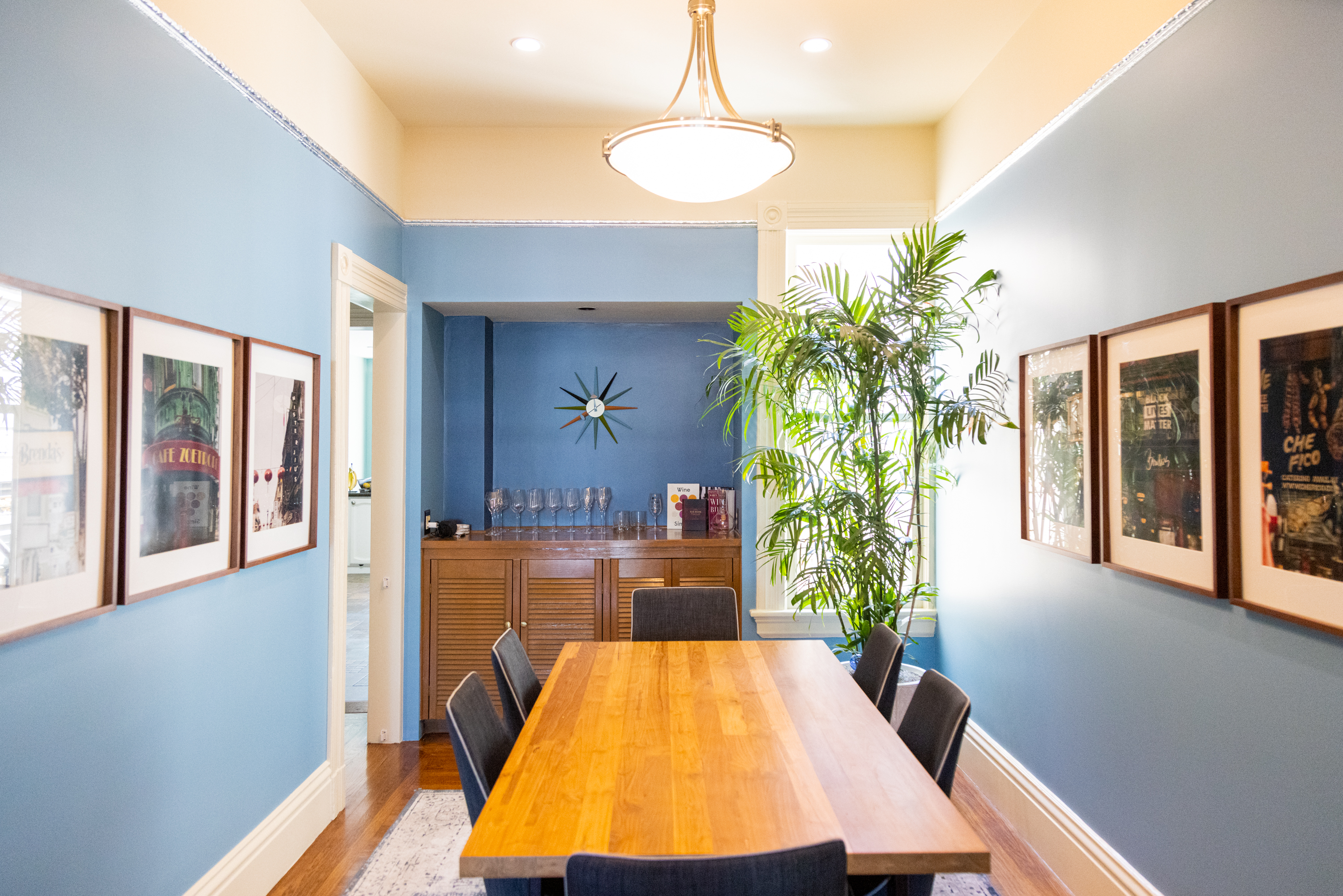 A dining room with a long wooden table, blue walls, framed posters, and a mid-century style clock.