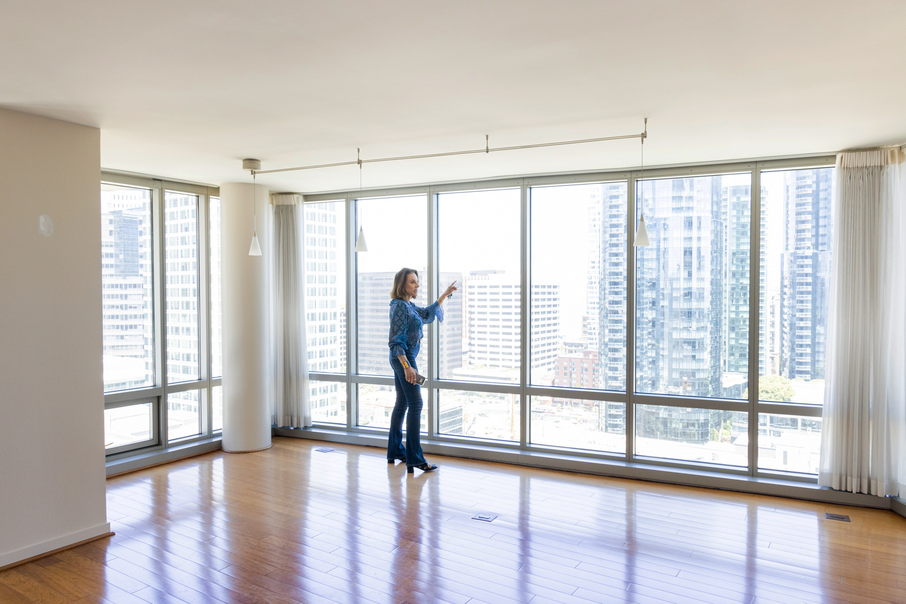 A woman stands in an empty, sunlit room with large floor-to-ceiling windows showcasing a cityscape of tall buildings, and she points towards the outside view.