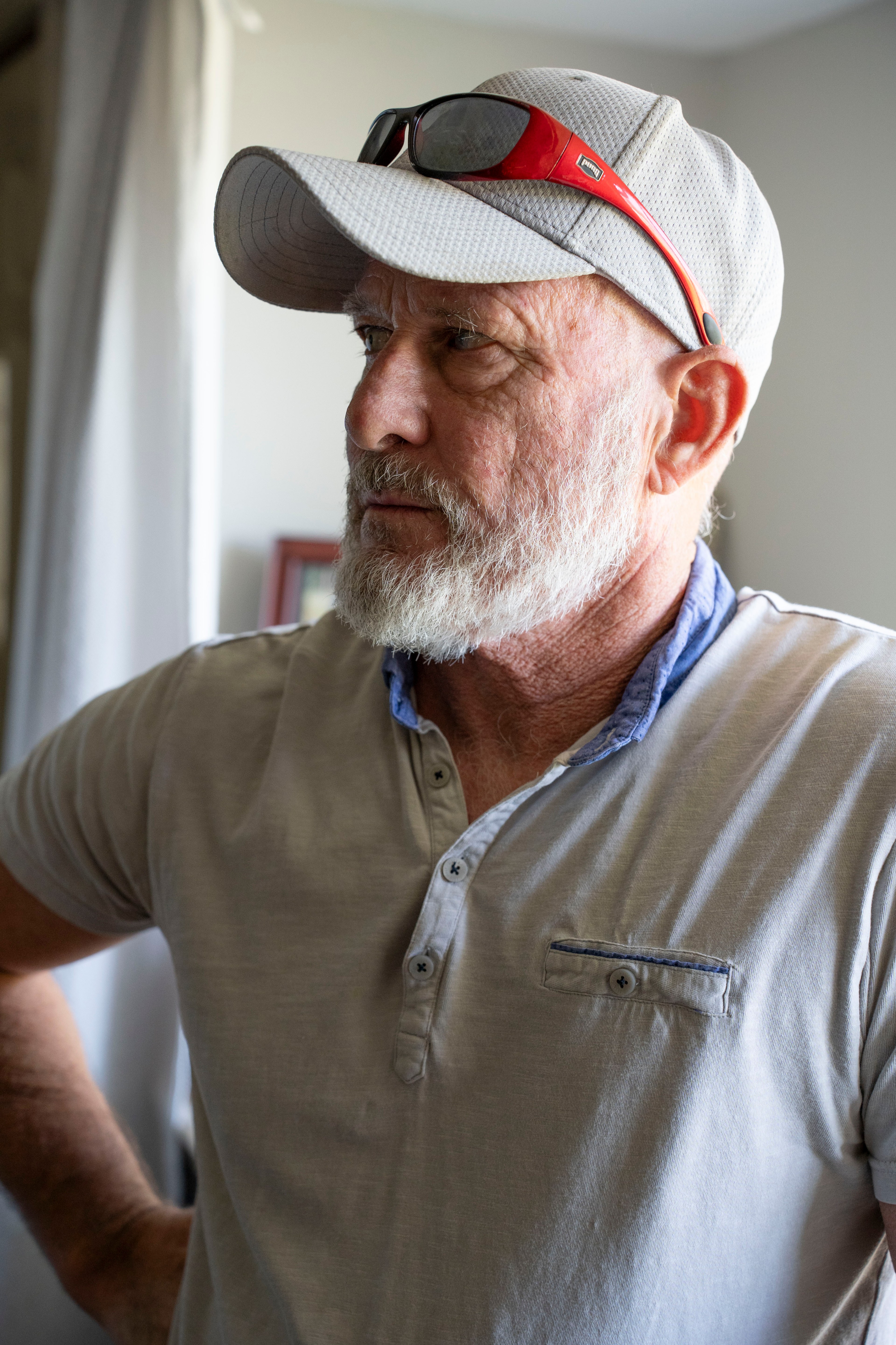 An older man with a beard wears a cap and sunglasses, looking to the side thoughtfully.