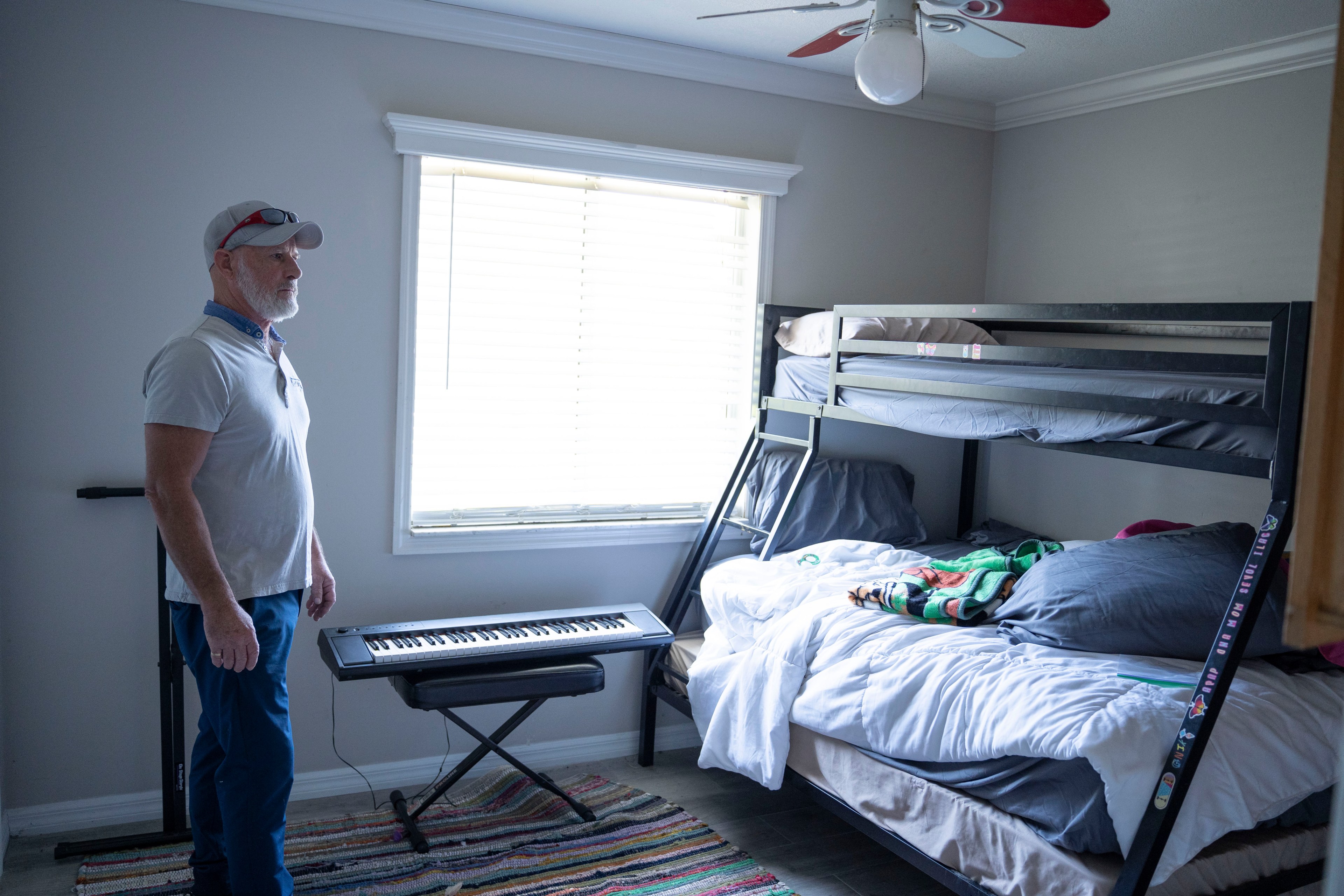 A man stands by a keyboard in a room with a bunk bed and a window with blinds.