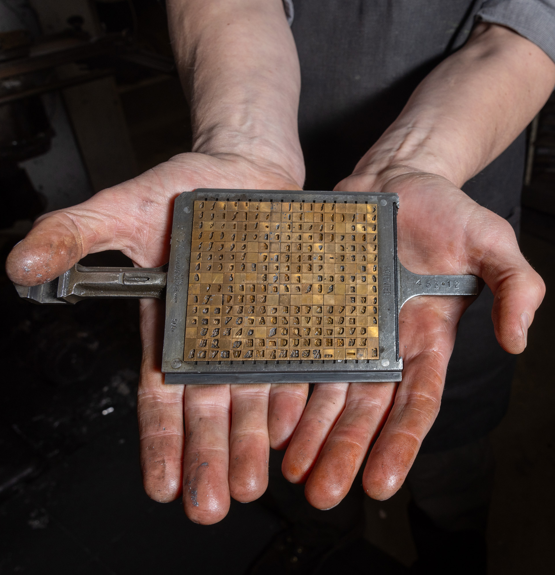 Dusty hands hold a large metal stamp with tiny, raised letters and numbers. It's used in printing or labeling.