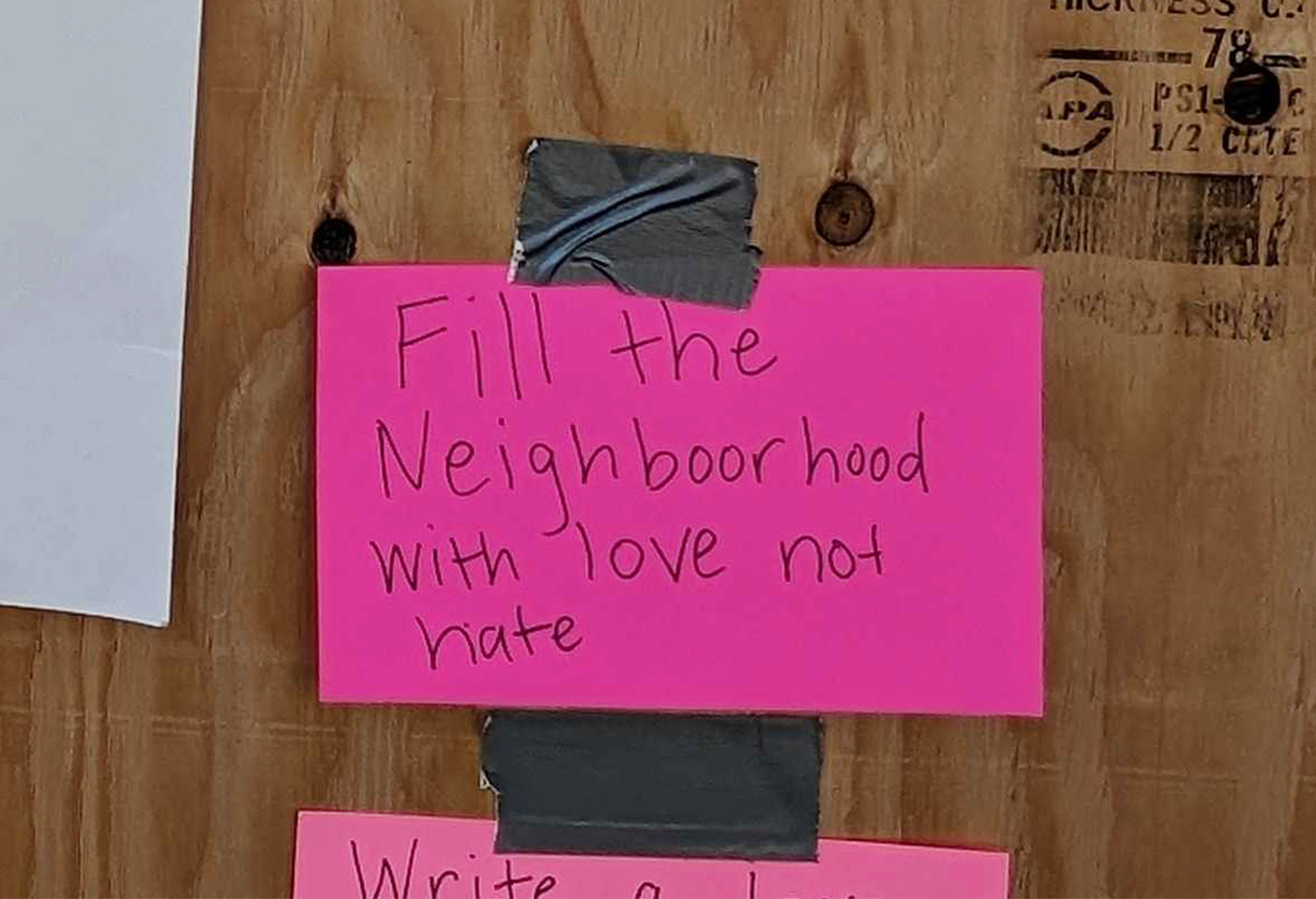 A neon pink card taped to a wooden board reads, &quot;Fill the Neighboorhood with love not hate,&quot; in handwritten text.