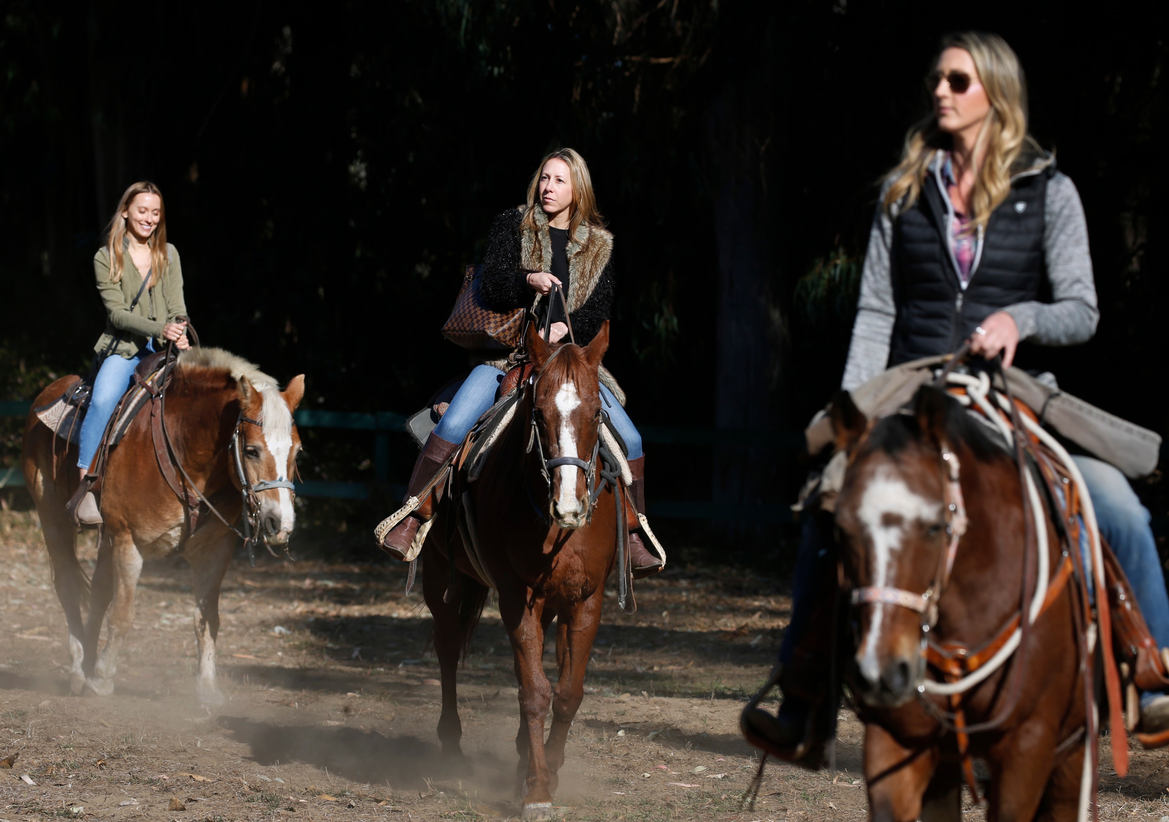 Three women ride horses on a sunny trail, one smiling, one serious, and one focused, casting shadows over a dusty path.