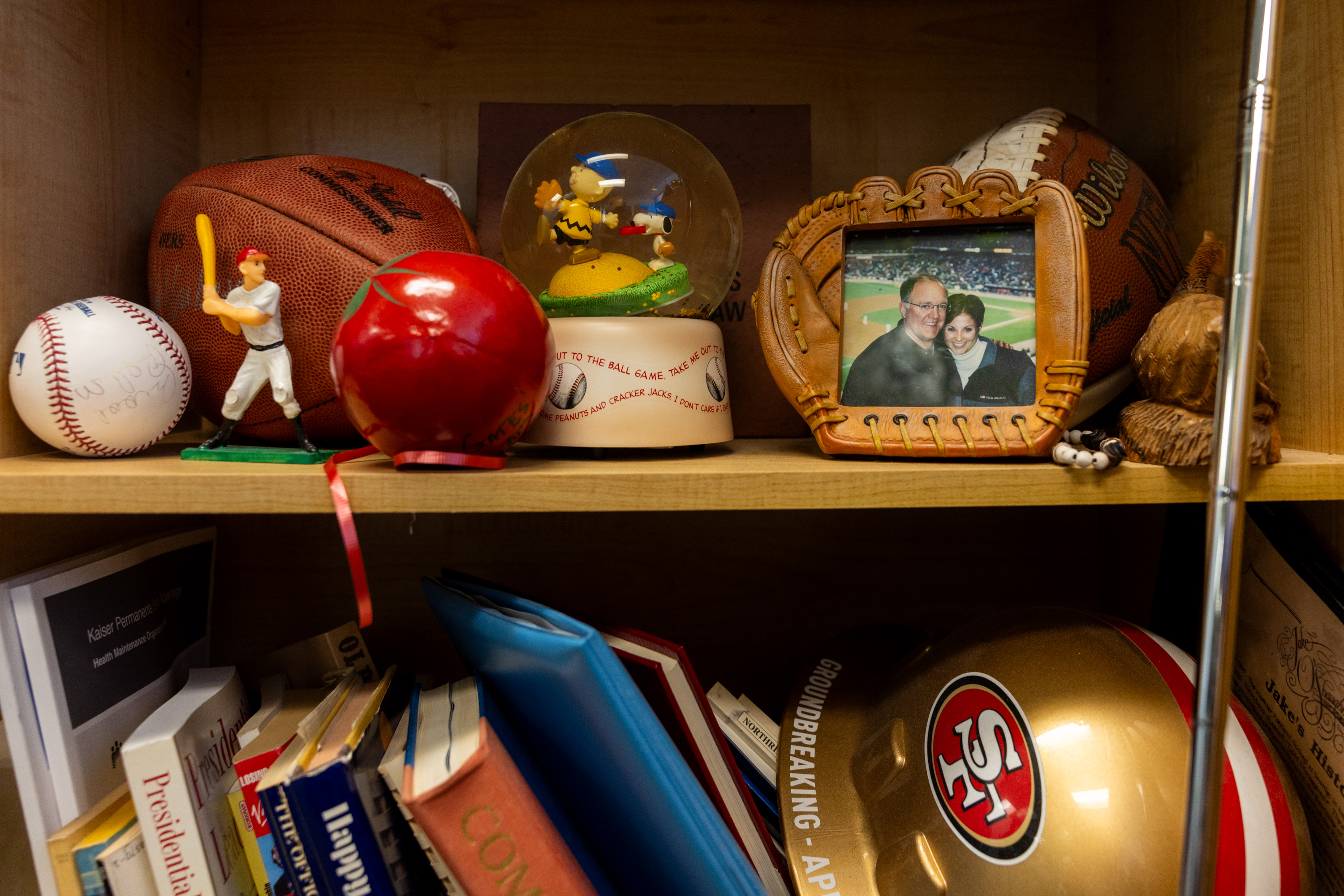 A shelf holds a basketball, baseball, figurine, snow globe, photo in a mitt frame, red apple, books, and a 49ers helmet. Other small objects are also present.