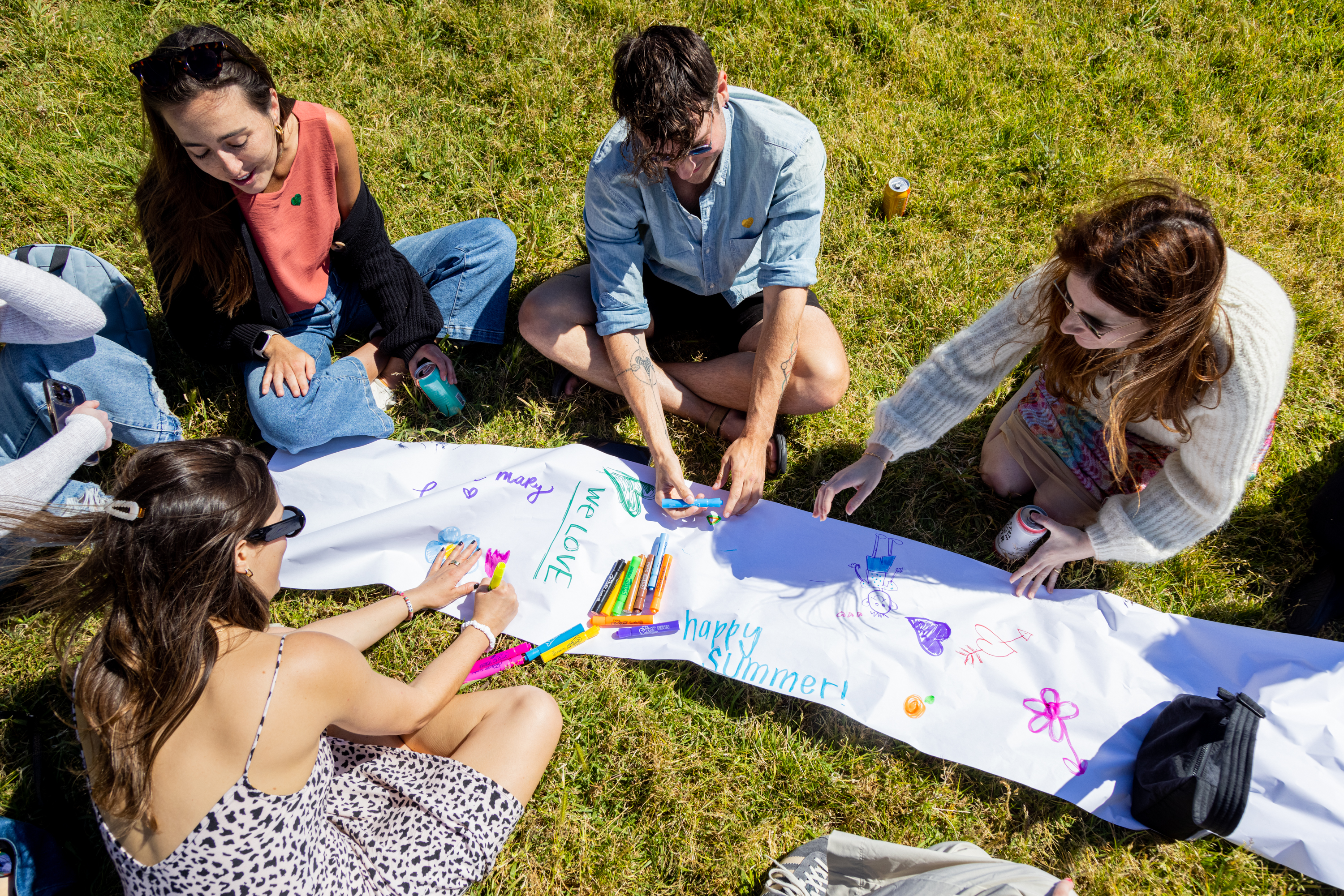 Four young people sit on grass, decorating a long banner with colorful markers, under a sunny sky. Their banner says &quot;happy summer!&quot; with artwork.