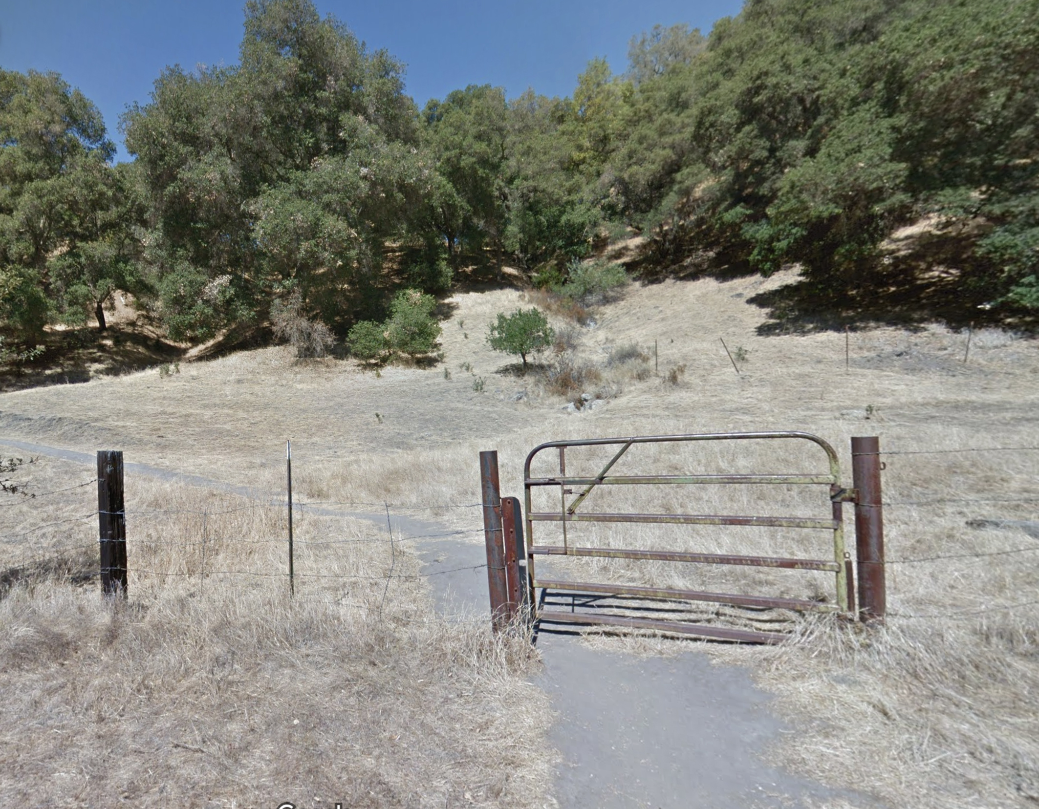 The image shows a rusty metal gate on a dirt path, with dry grass and scattered bushes around. Behind the gate are several trees on a sloping hill under a clear blue sky.
