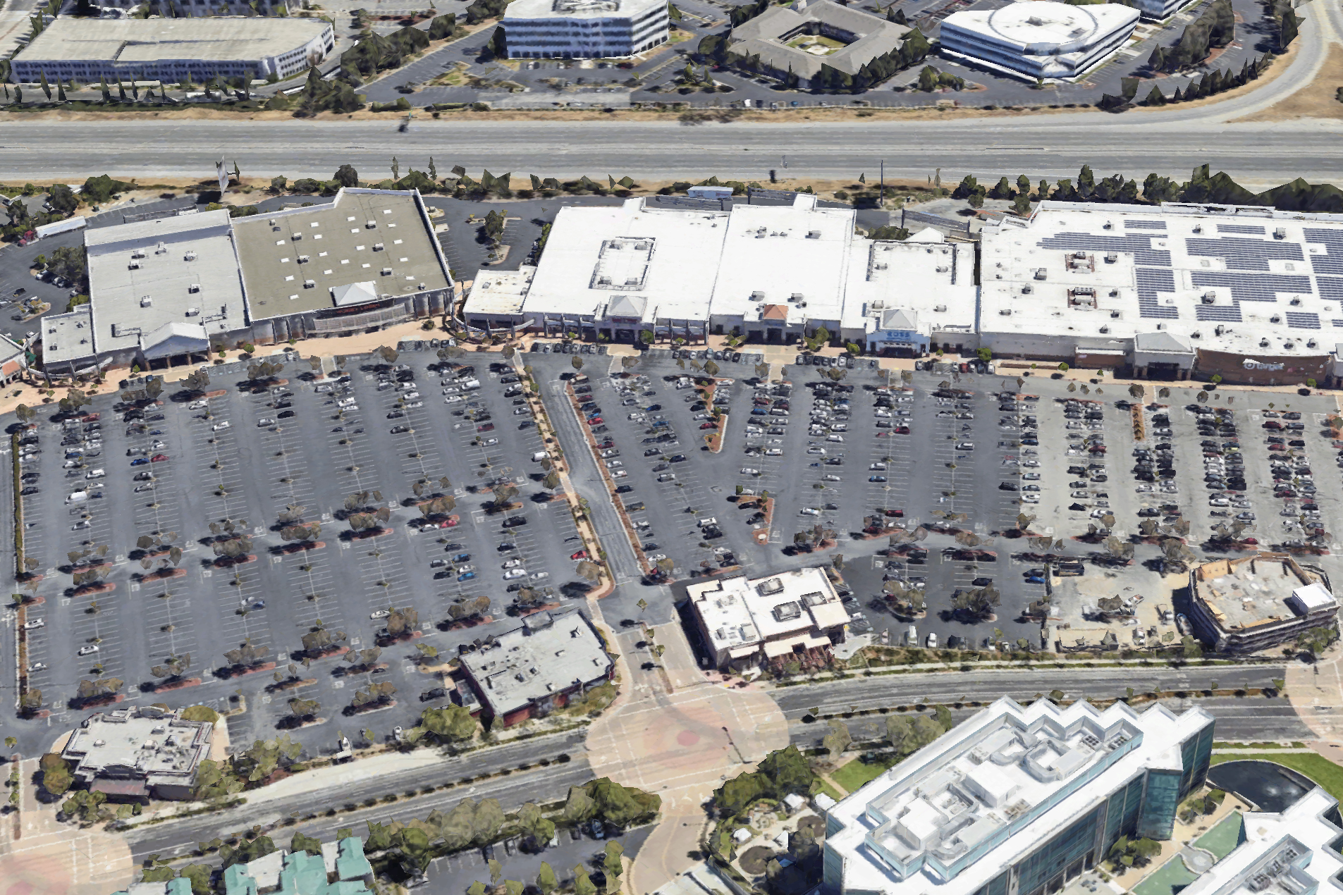 The image shows an aerial view of a large parking lot in front of several big retail stores, with many cars parked. Surrounding roads and buildings are visible.