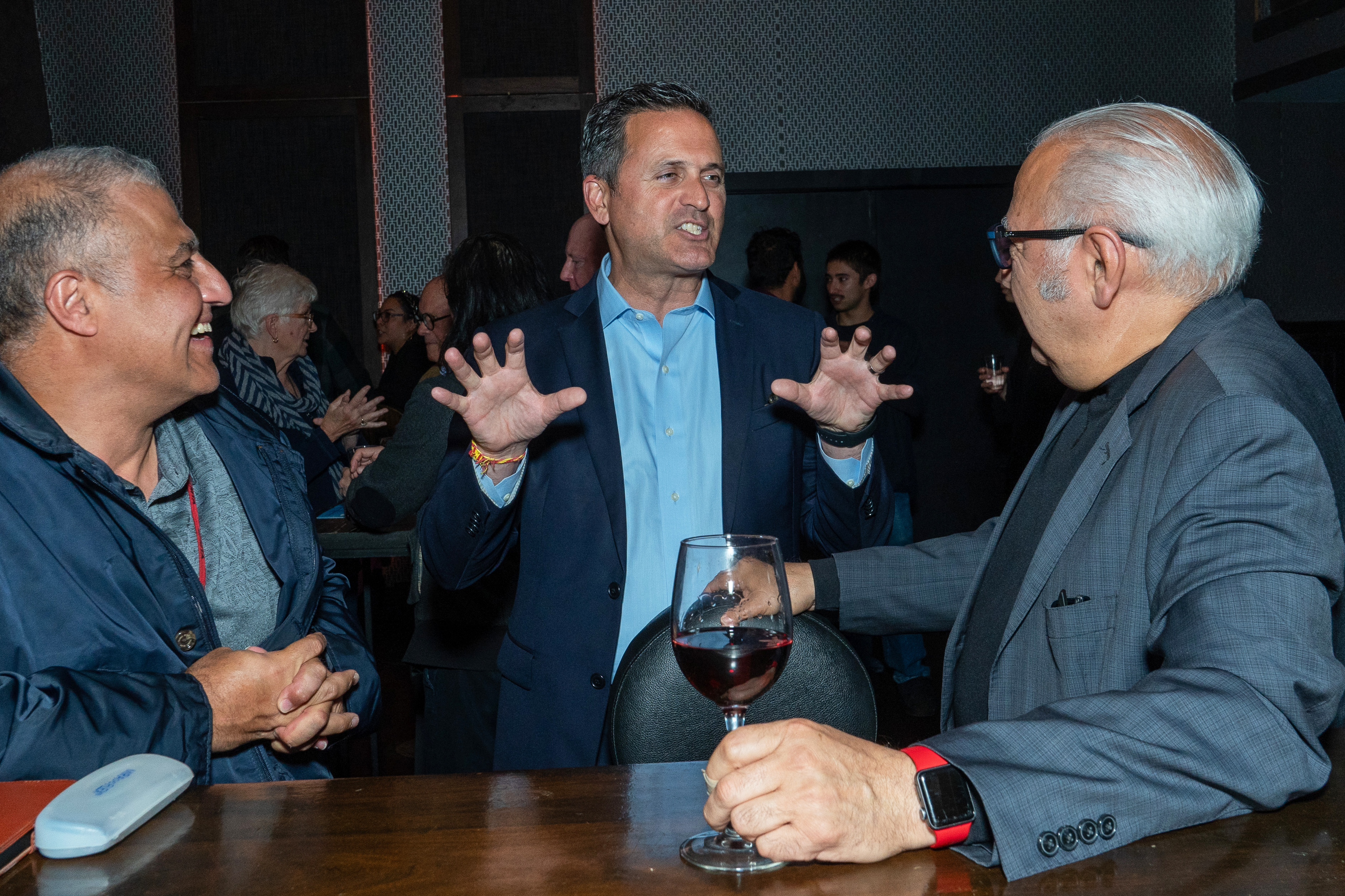 Three men share a conversation at a social event, one gesturing animatedly while holding a wine glass. They are dressed in business casual attire.