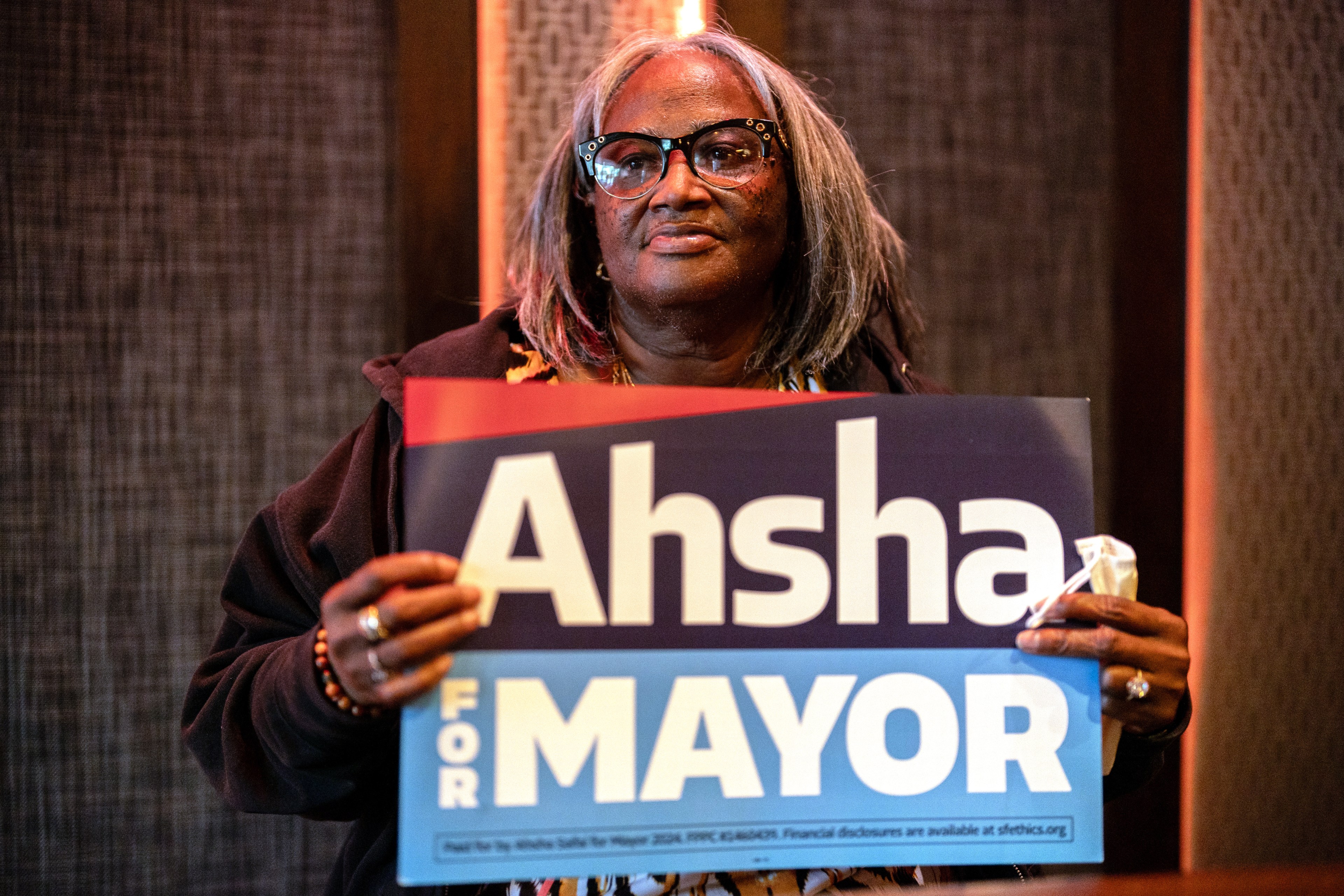An individual holds a campaign sign reading &quot;Ahsha for Mayor&quot;, expressing a serious demeanor. They wear glasses and a brown sweater.