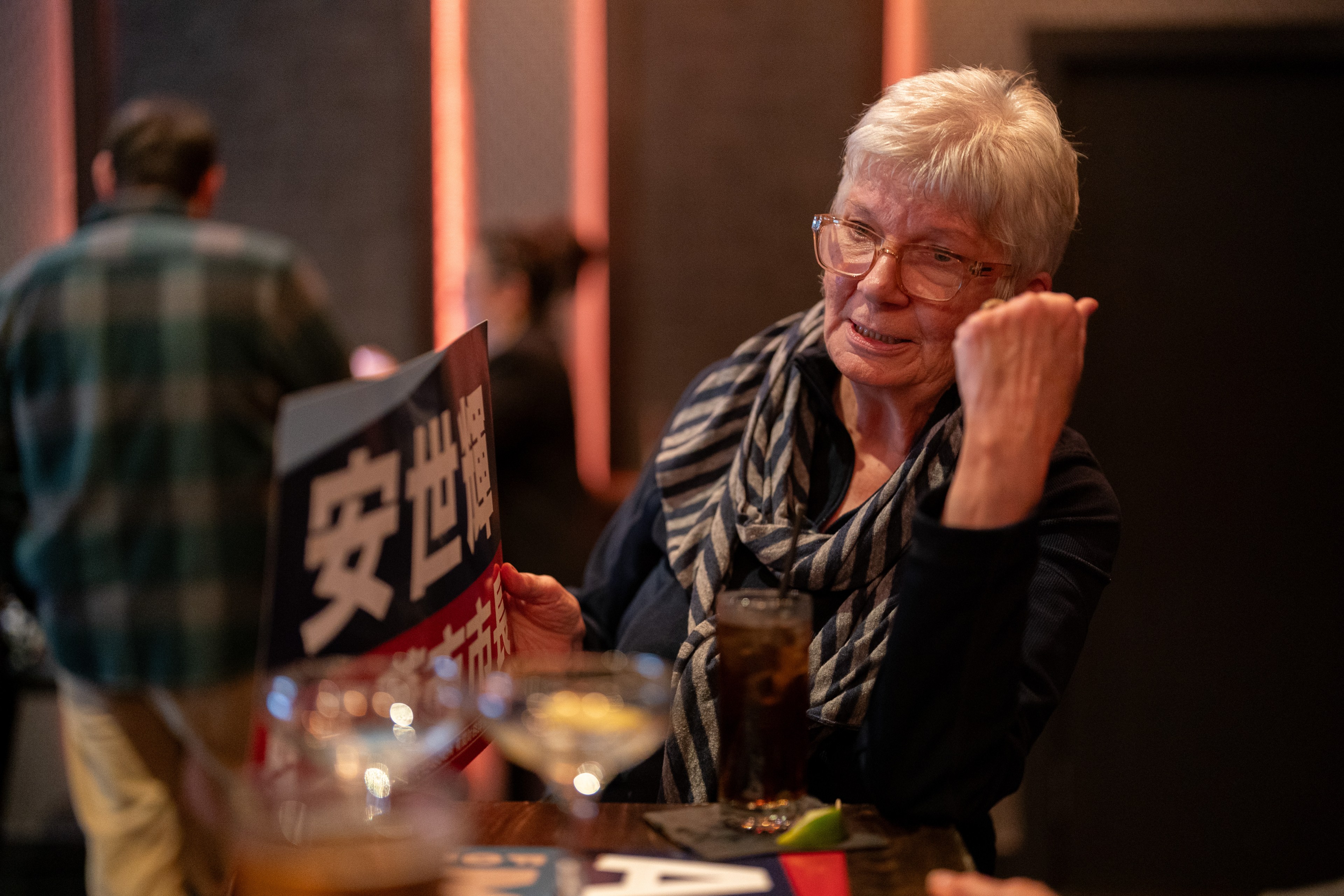 An elderly woman in a bar holds a menu with Japanese characters, smiling while interacting with someone off-camera; drinks and soft lighting in the background.