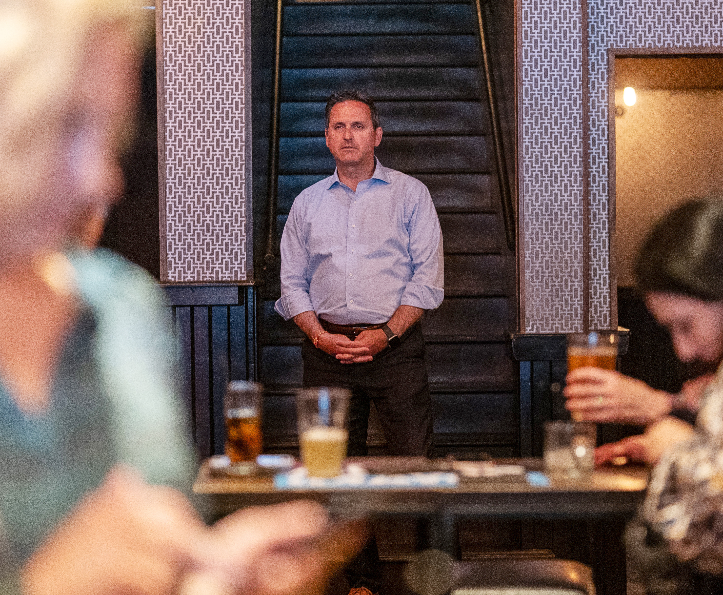 A man in a blue shirt stands attentively in a dimly lit bar, surrounded by blurred figures focused on their drinks and conversations.