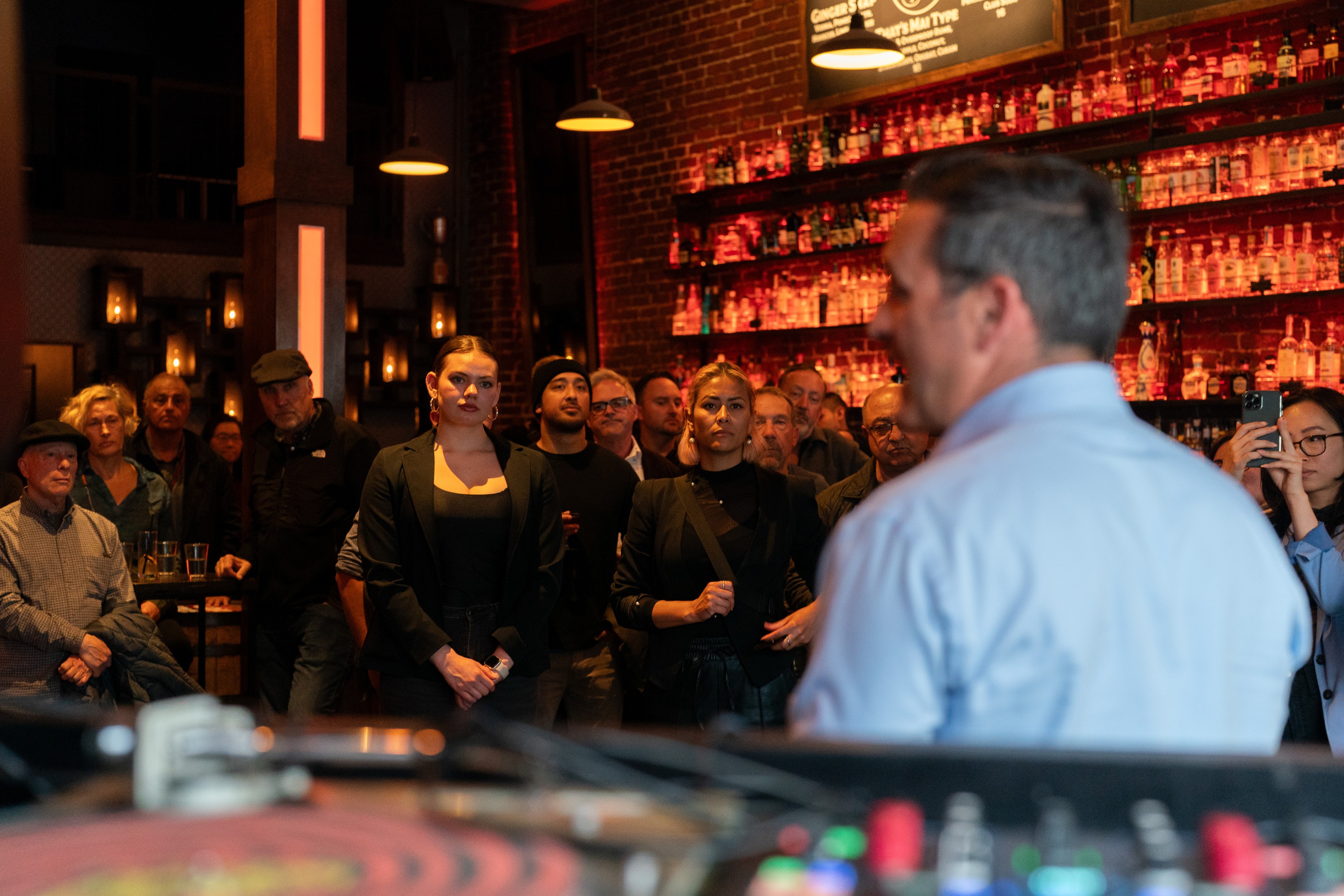 A group listens attentively to a speaker at a bar, illuminated by ambient lighting and a backlit shelf of bottles.
