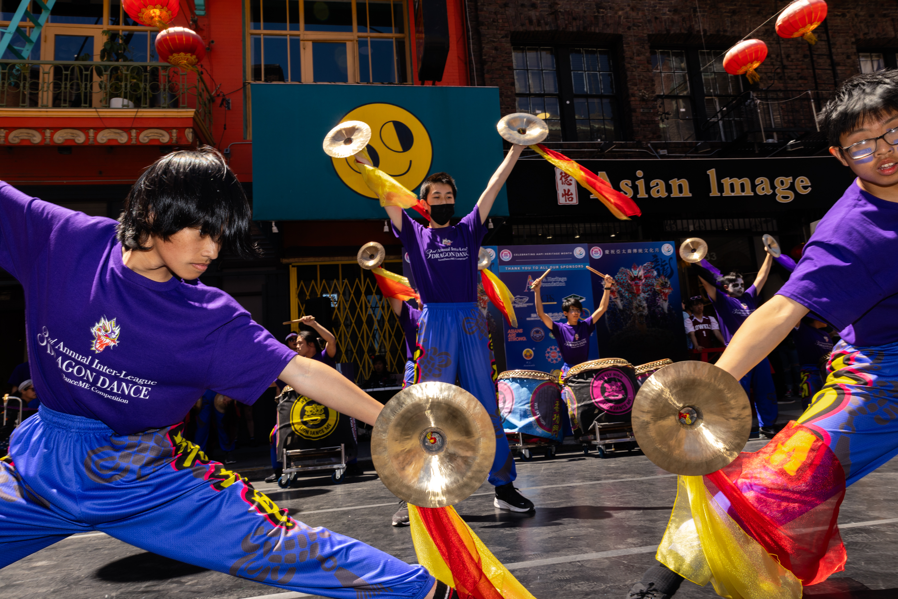 Performers in purple outfits with cymbals dance in a vibrant street celebration, backed by smiling sun decor.