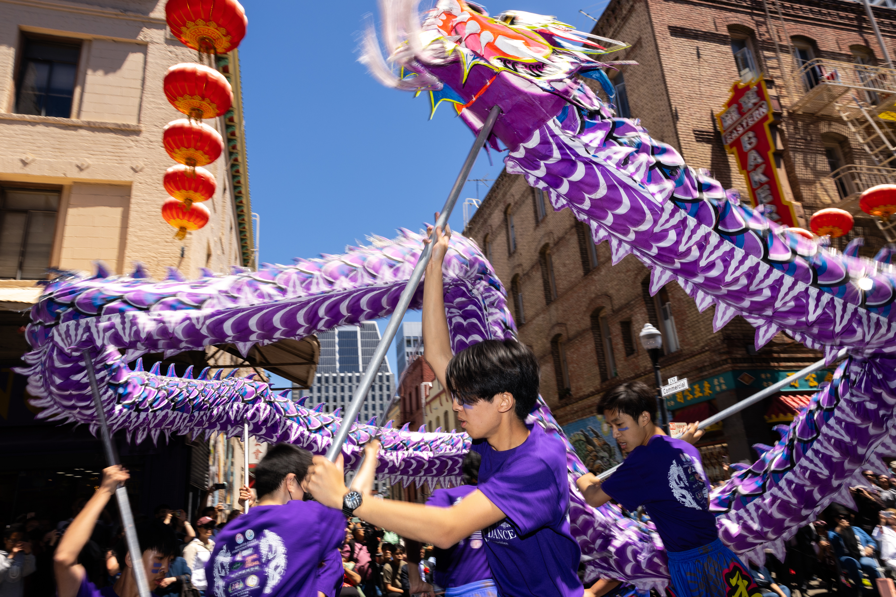 People perform a dragon dance under red lanterns in a sunny street parade.