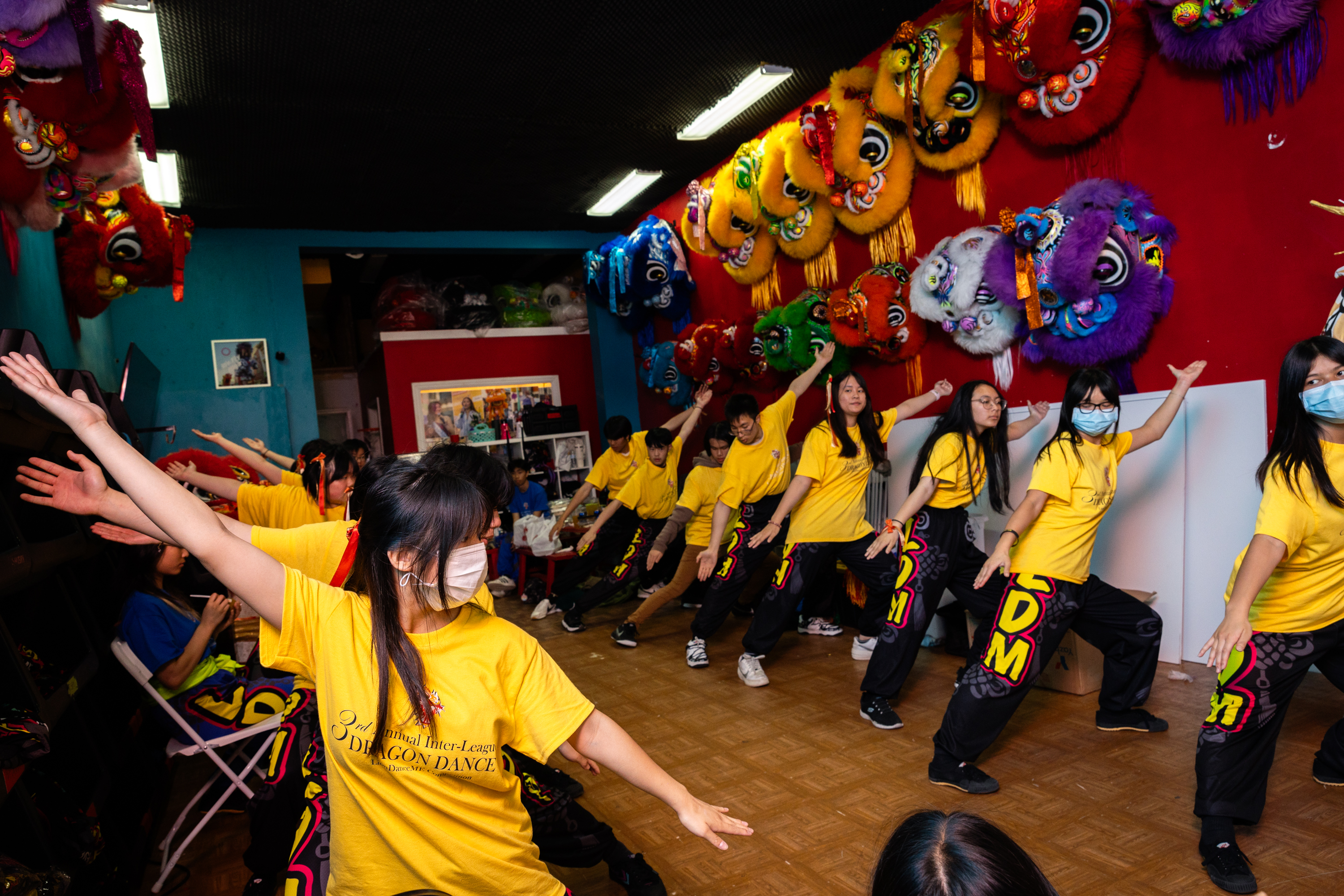People in yellow shirts and masks are practicing a dance, with colorful lion dance masks decorating the wall behind them.
