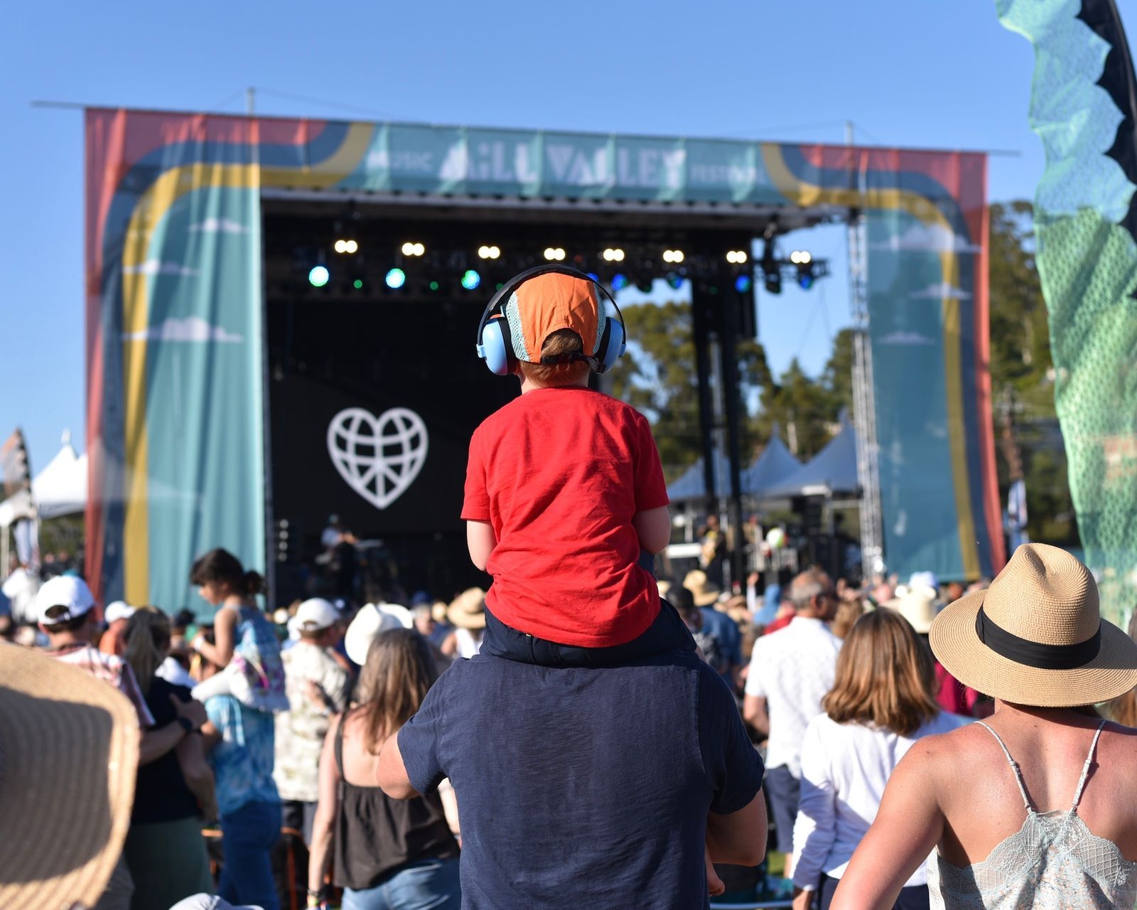 A child in a red shirt and headphones sits on an adult's shoulders at an outdoor festival, facing a stage with a heart logo.