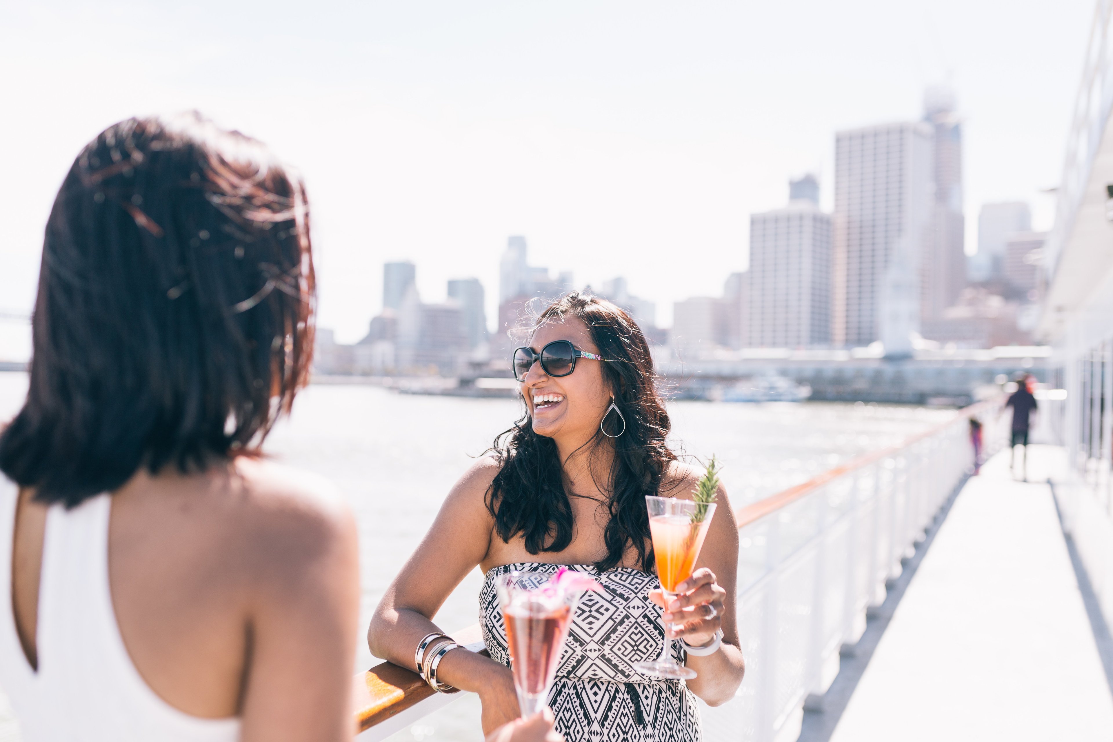 Two women on a boat with drinks, one laughing with a city skyline in the background.