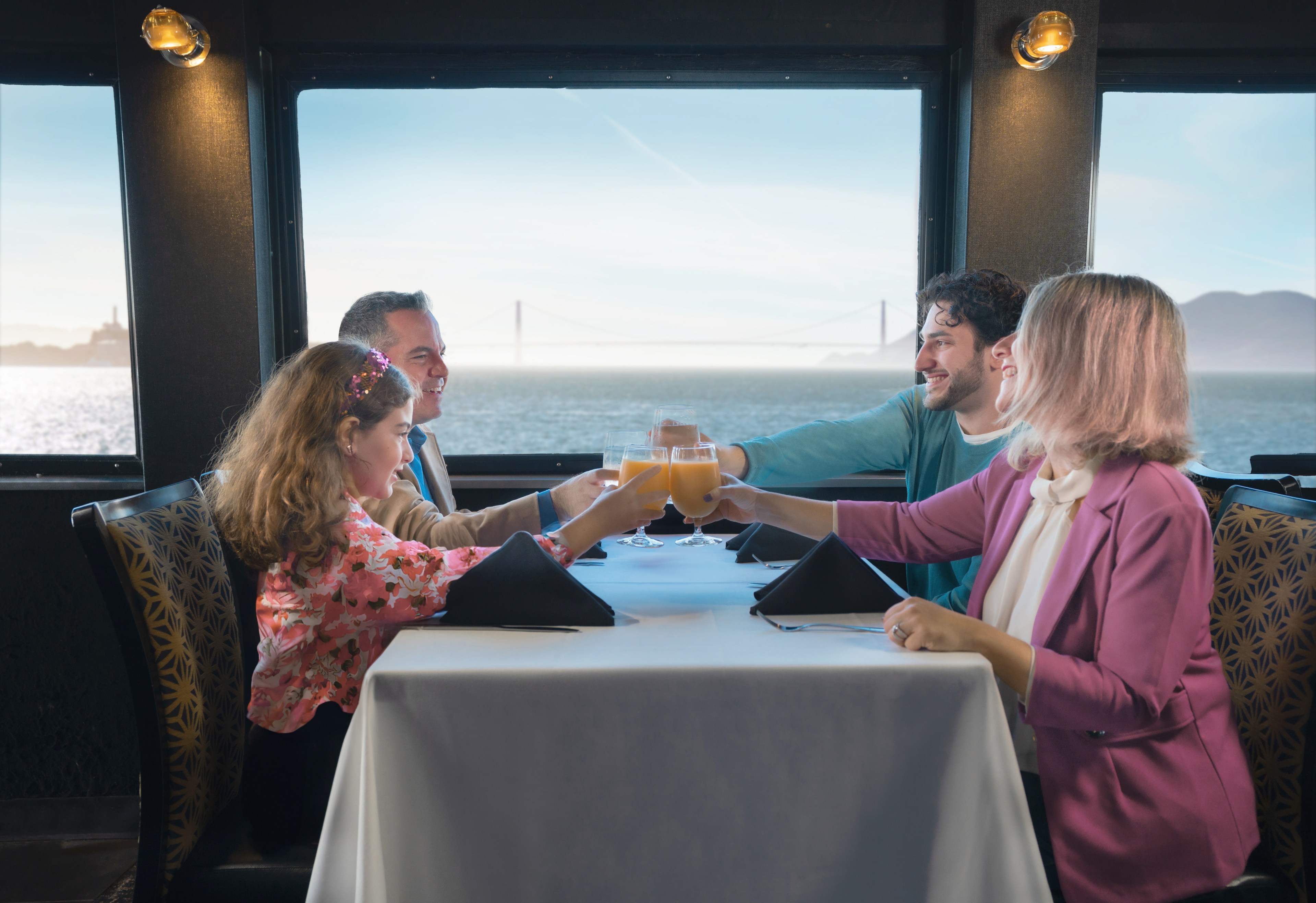 Four people are toasting drinks at a table by a window with a bridge and water outside.