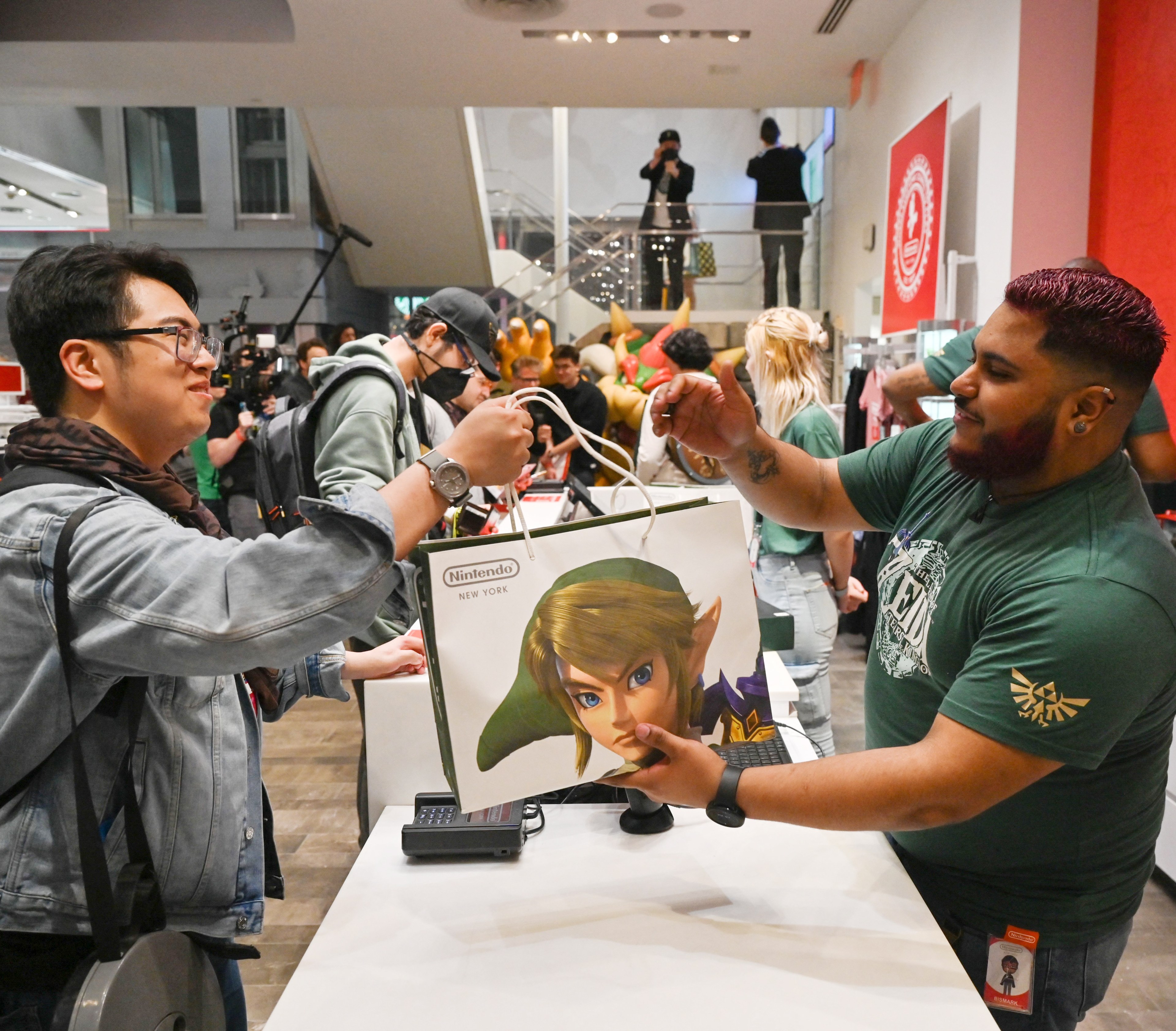 A man hands a Nintendo shopping bag featuring Link from The Legend of Zelda to a customer in a busy store with people and gaming merchandise in the background.