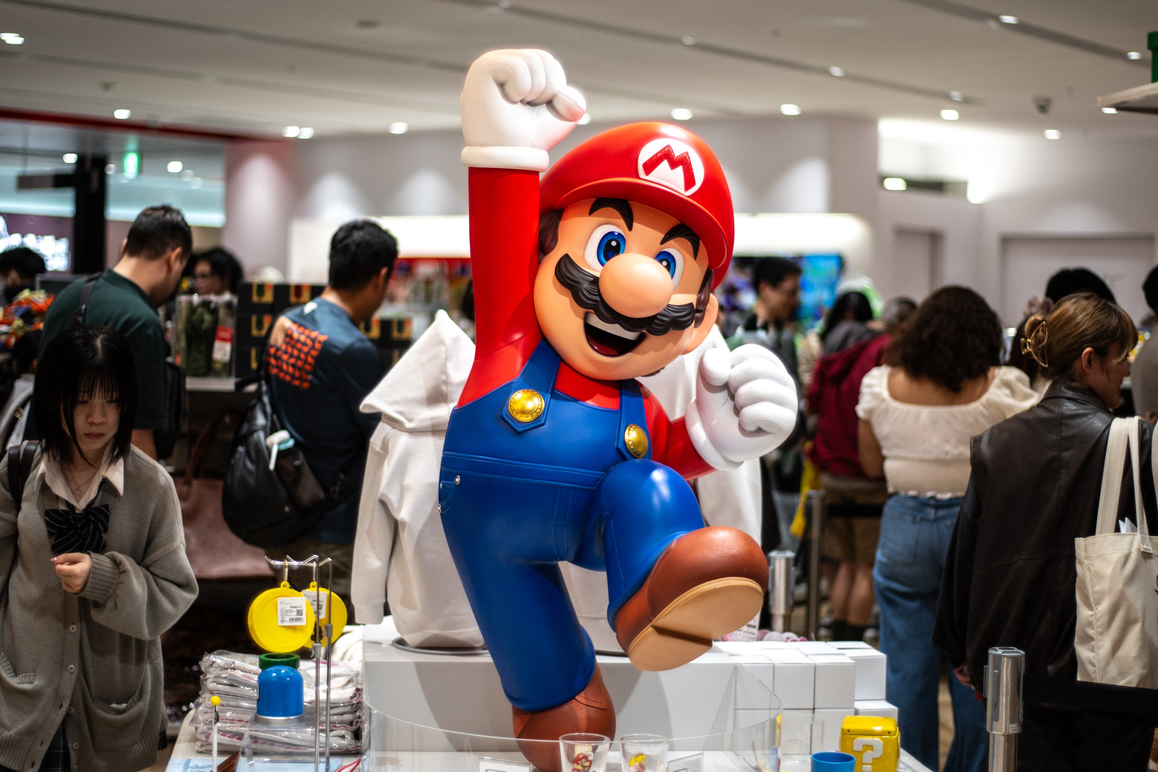 A large Mario statue is center stage, surrounded by bustling people in a store. Mario is in his distinctive red hat and blue overalls, striking a joyful pose.