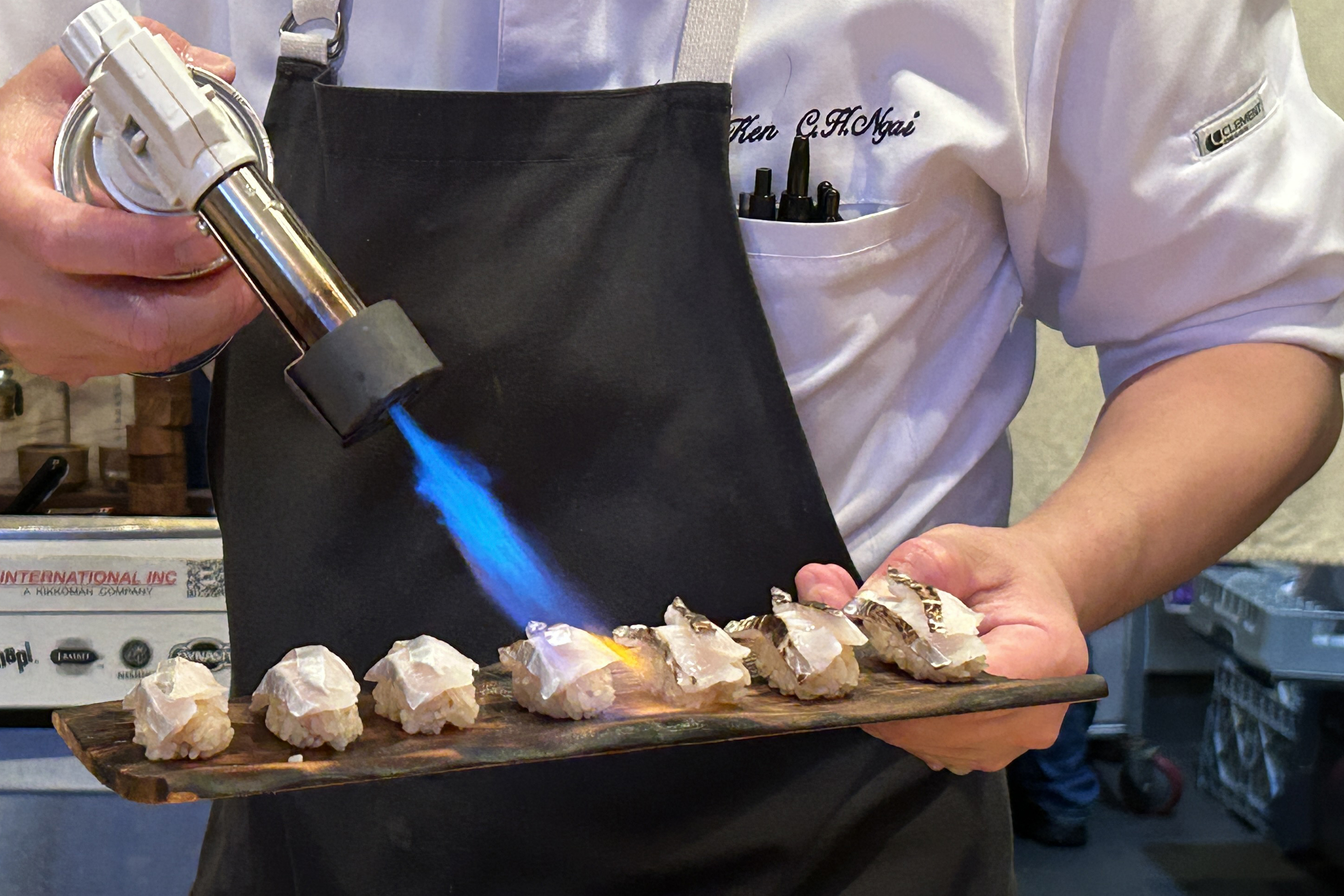 A chef uses a blowtorch to sear a row of sushi pieces arranged on a wooden tray, each topped with a thin slice of fish.