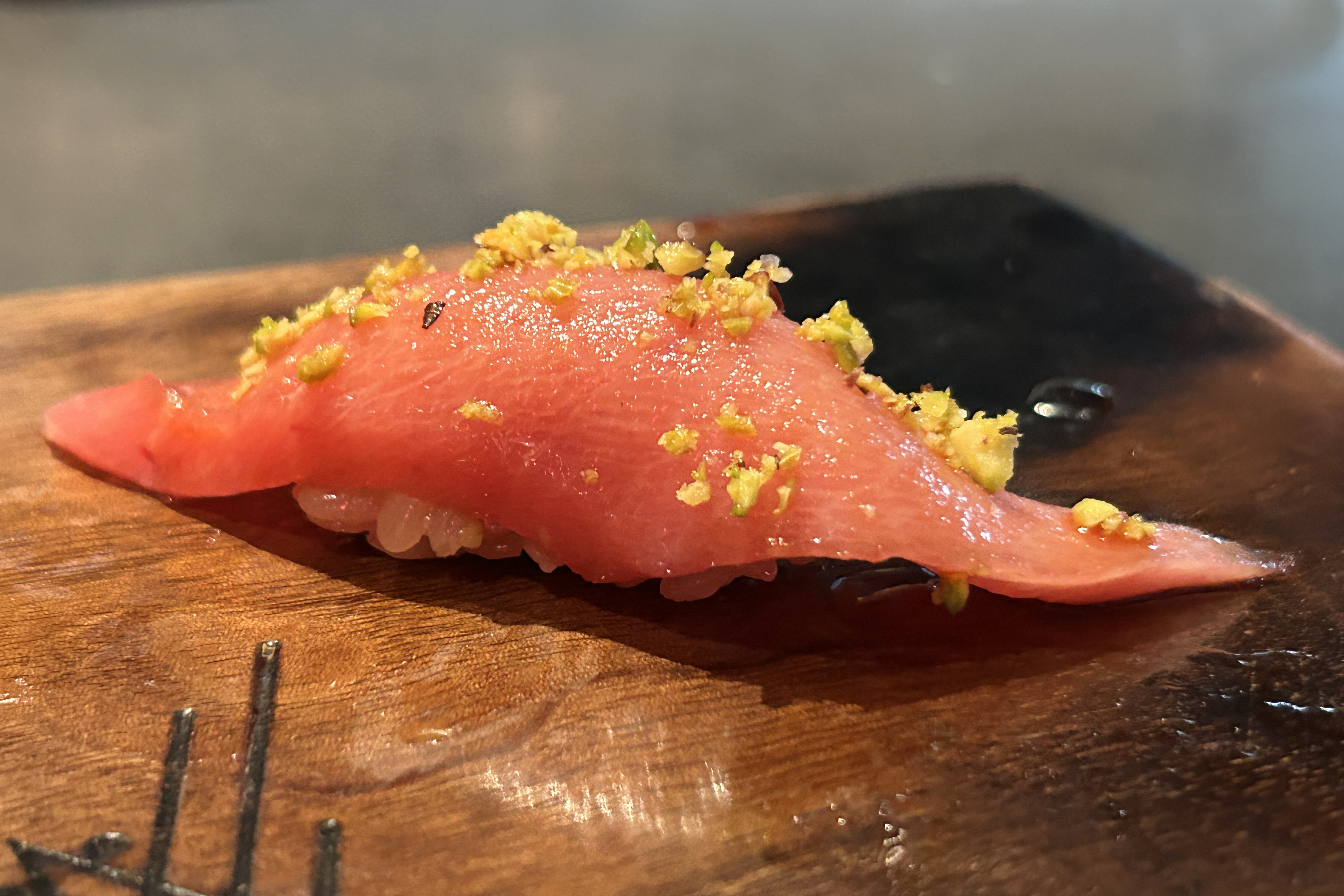 A piece of sushi with a slice of raw fish, likely tuna, is placed on a wooden surface, garnished with yellowish seasoning.