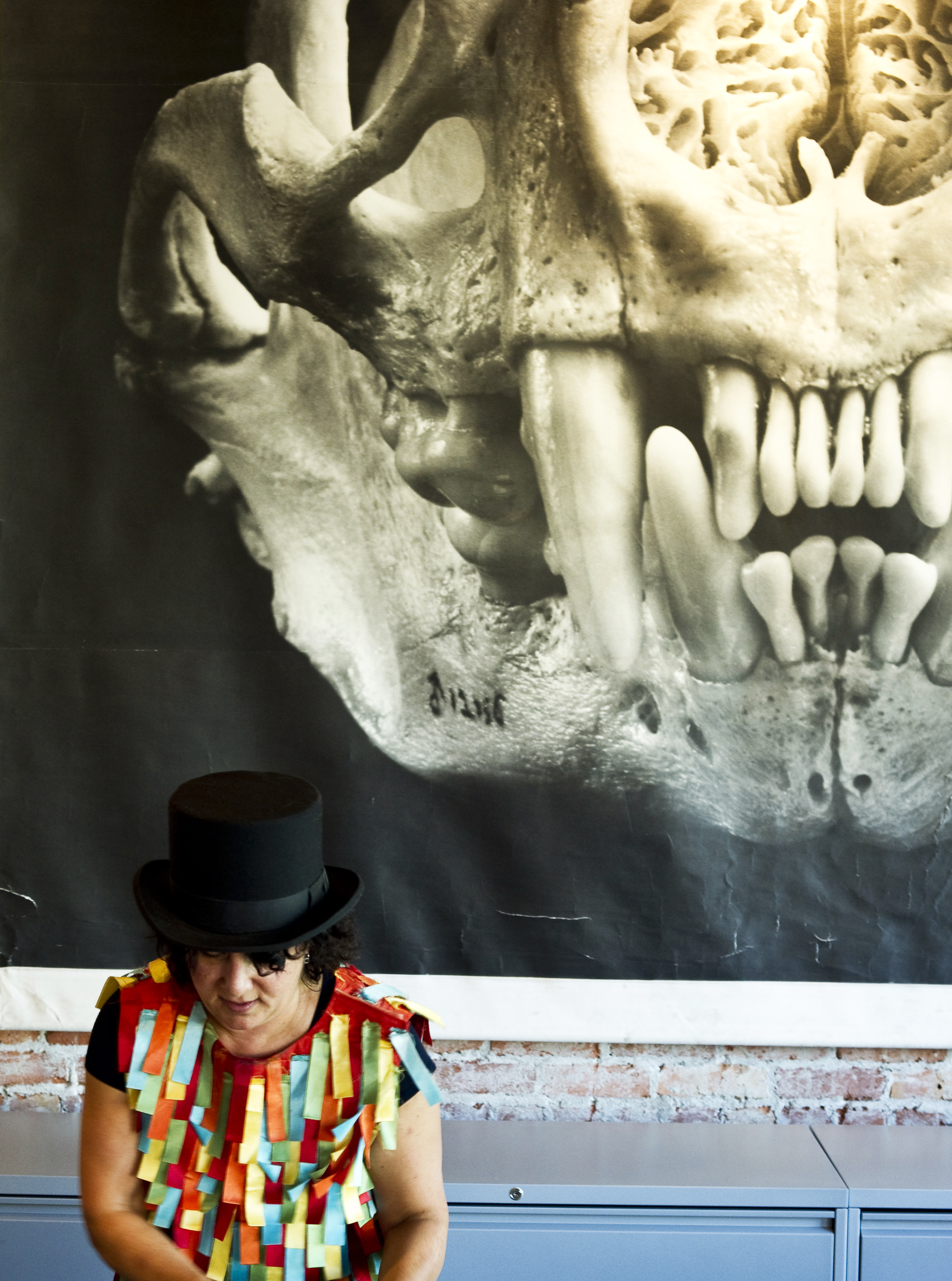 A person with a black top hat and colorful, ribbon-like attire stands in front of a large, black-and-white skull image pinned on a wall.