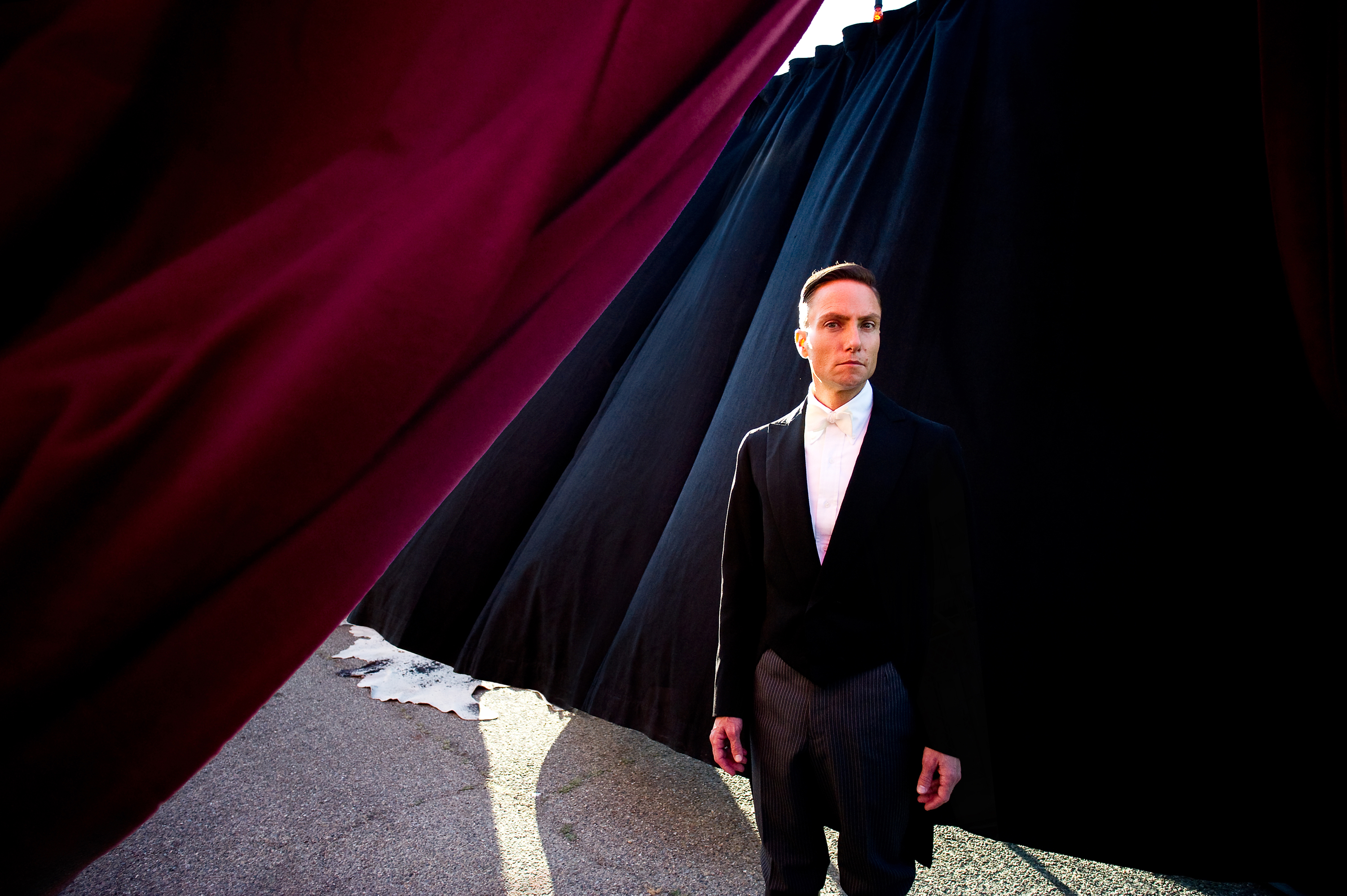 A man in a black tuxedo with a white shirt stands between large red and black curtains, with part of the ground visible beneath them. The setting appears formal.