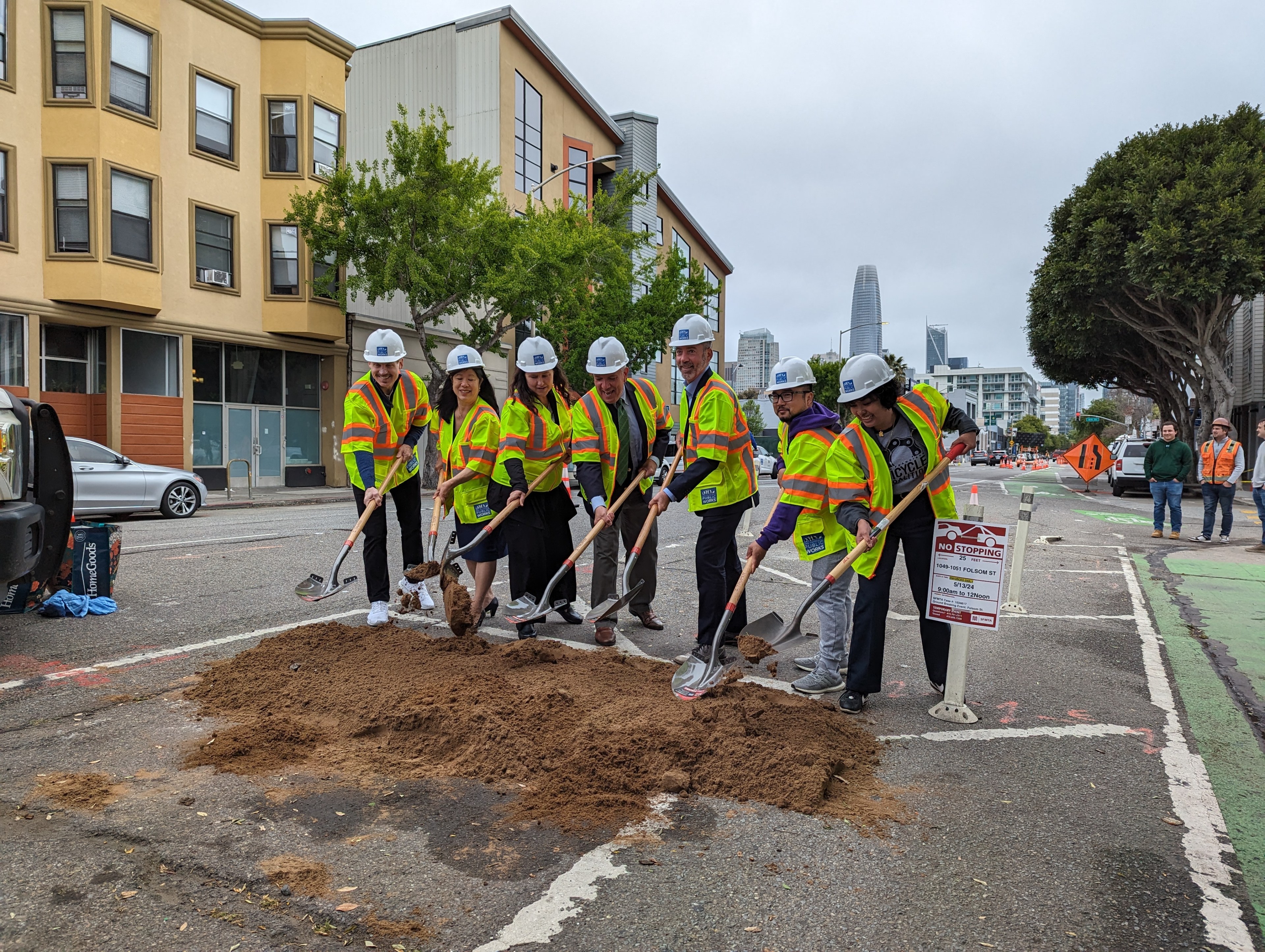 A group of people wearing hard hats shovel dirt on a city street.