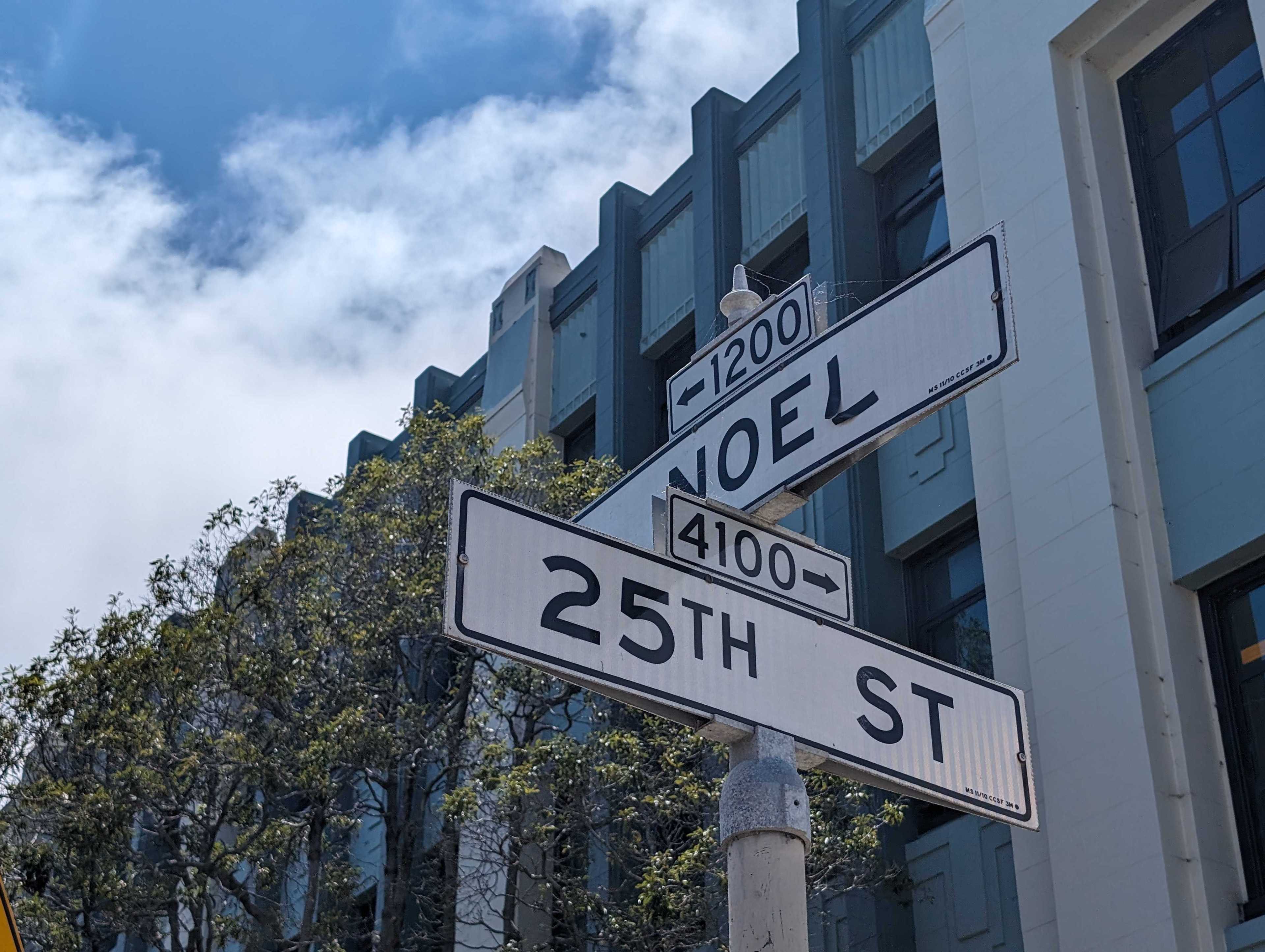 Street signs for "NOEL" and "25TH ST" are mounted on a post in front of a modern building with trees and a blue sky.
